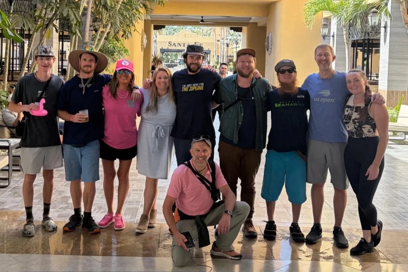 Group of eleven people standing together indoors, smiling for a photo. They are casually dressed, some wearing hats, sunglasses, and carrying bags or drinks. The setting appears to be a lobby or entrance area with plants and a yellow archway in the background.