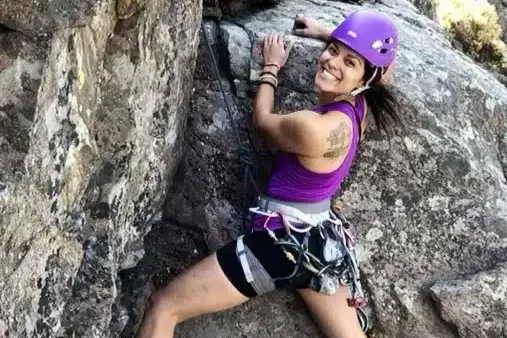 Woman wearing a purple helmet and tank top rock climbing on a rocky surface.