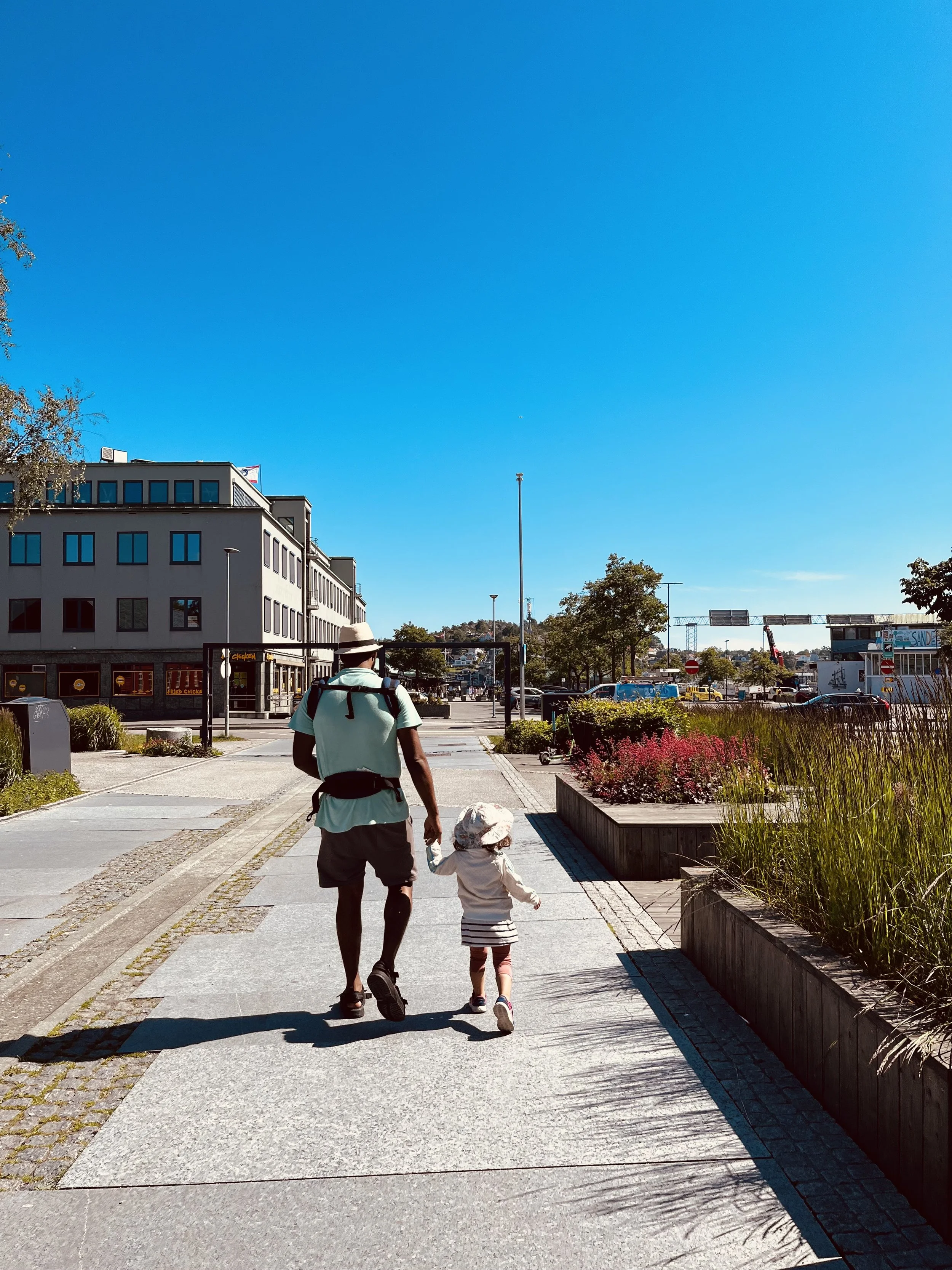 En voksen og et barn går hånd i hånd langs en bysgate med blomsterbeds og bygninger på begge sider, under en klar blå himmel.