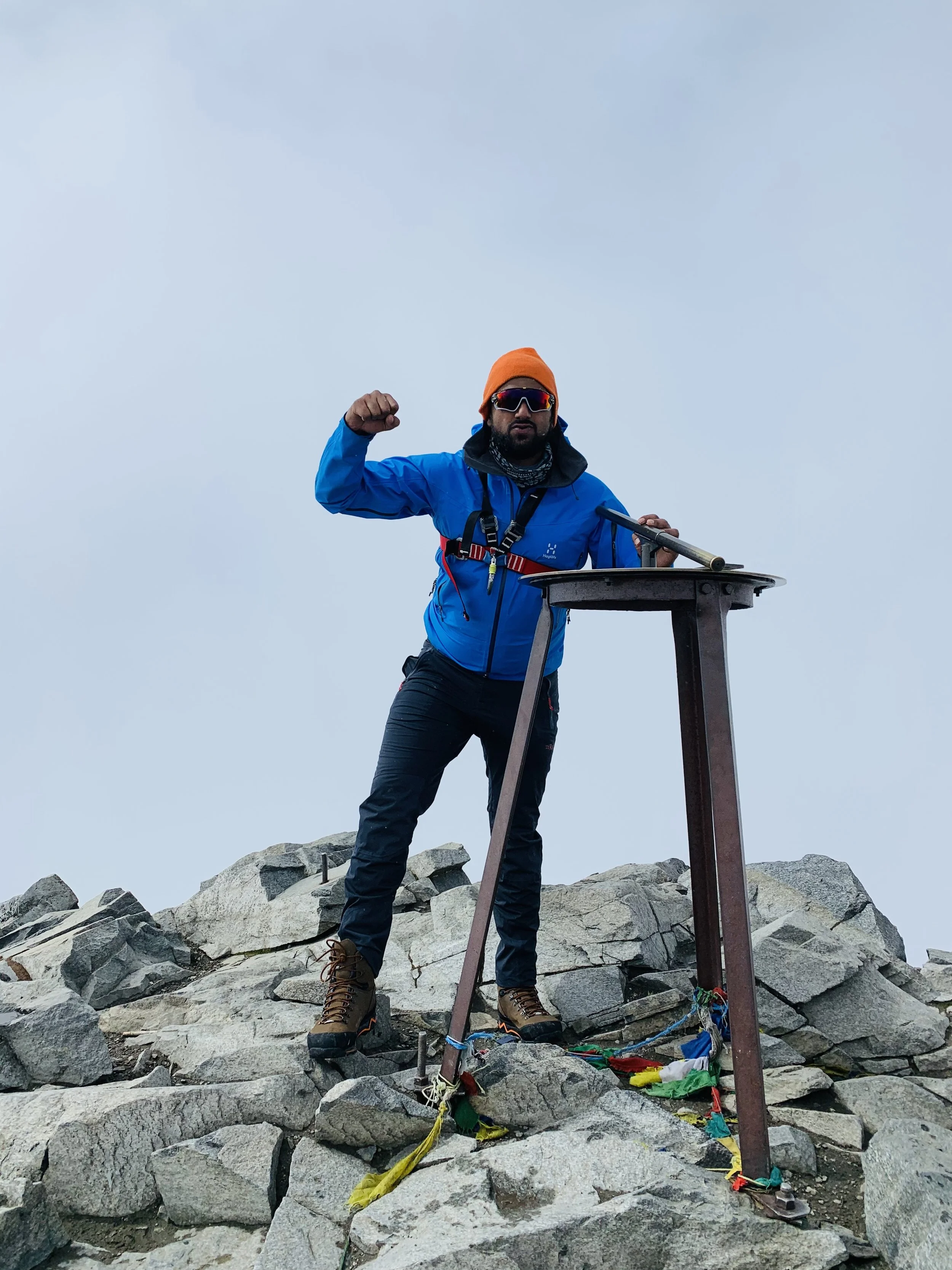 En mann står på en fjelltopp med steiner og fjell i bakgrunnen, iført fjellklatringsutstyr og løfter høyre arm i en seiersposisjon.