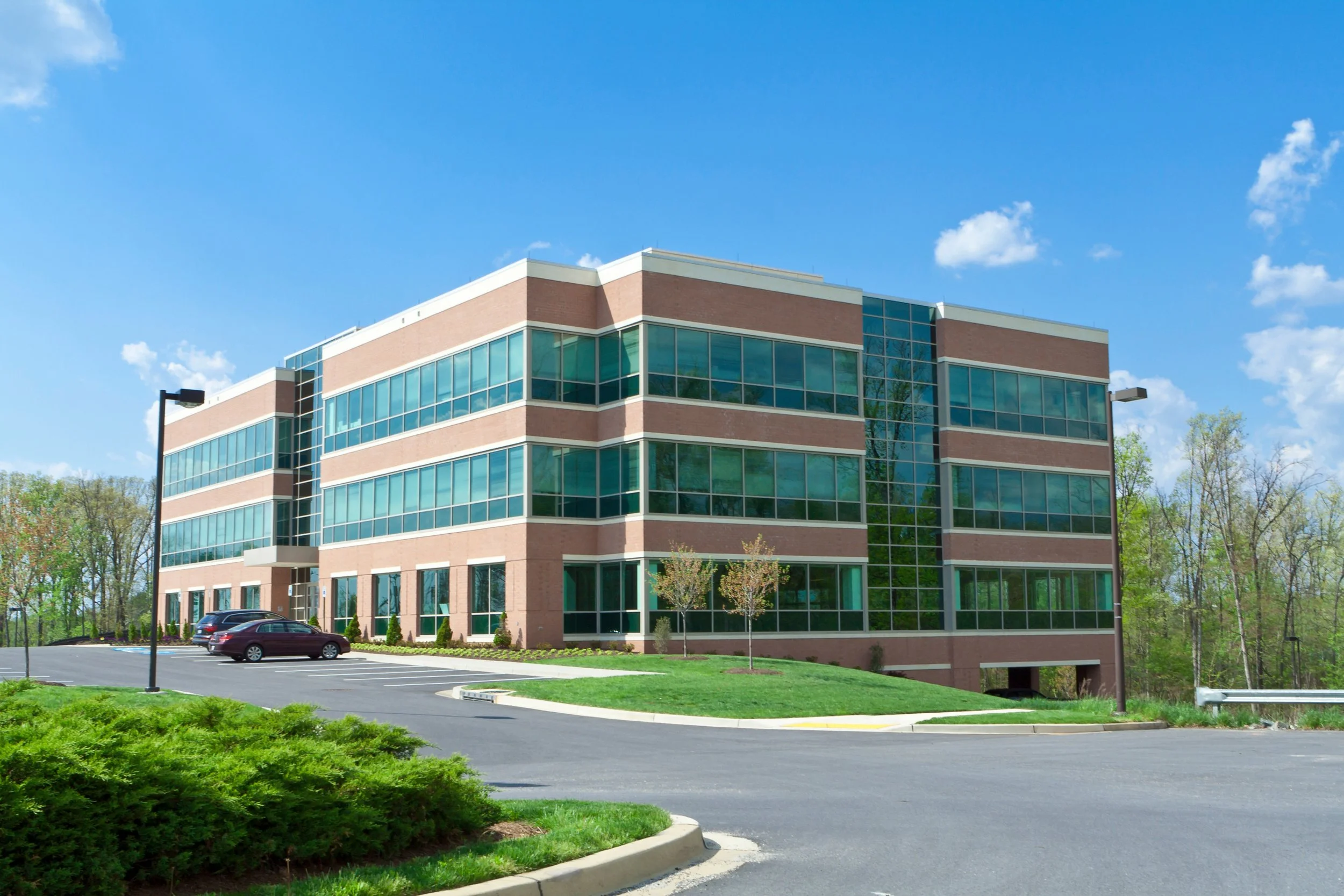 A modern four-story office building with large glass windows, surrounded by a parking lot and green landscaping under a clear blue sky.