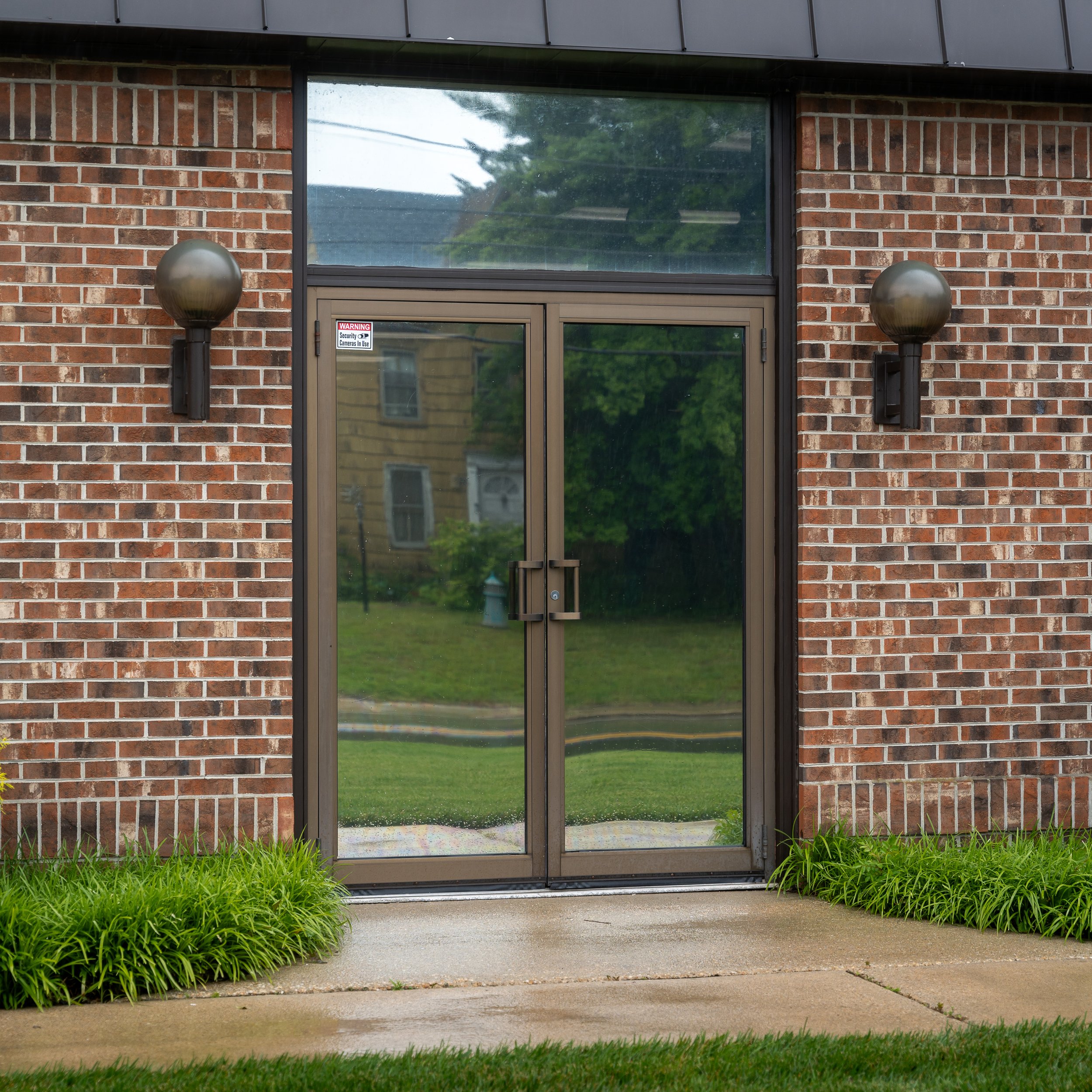 Glass double doors with metal frame, flanked by two outdoor wall sconces on a brick building, with green grass and plants in front. Tinted with Dual Reflective window tint