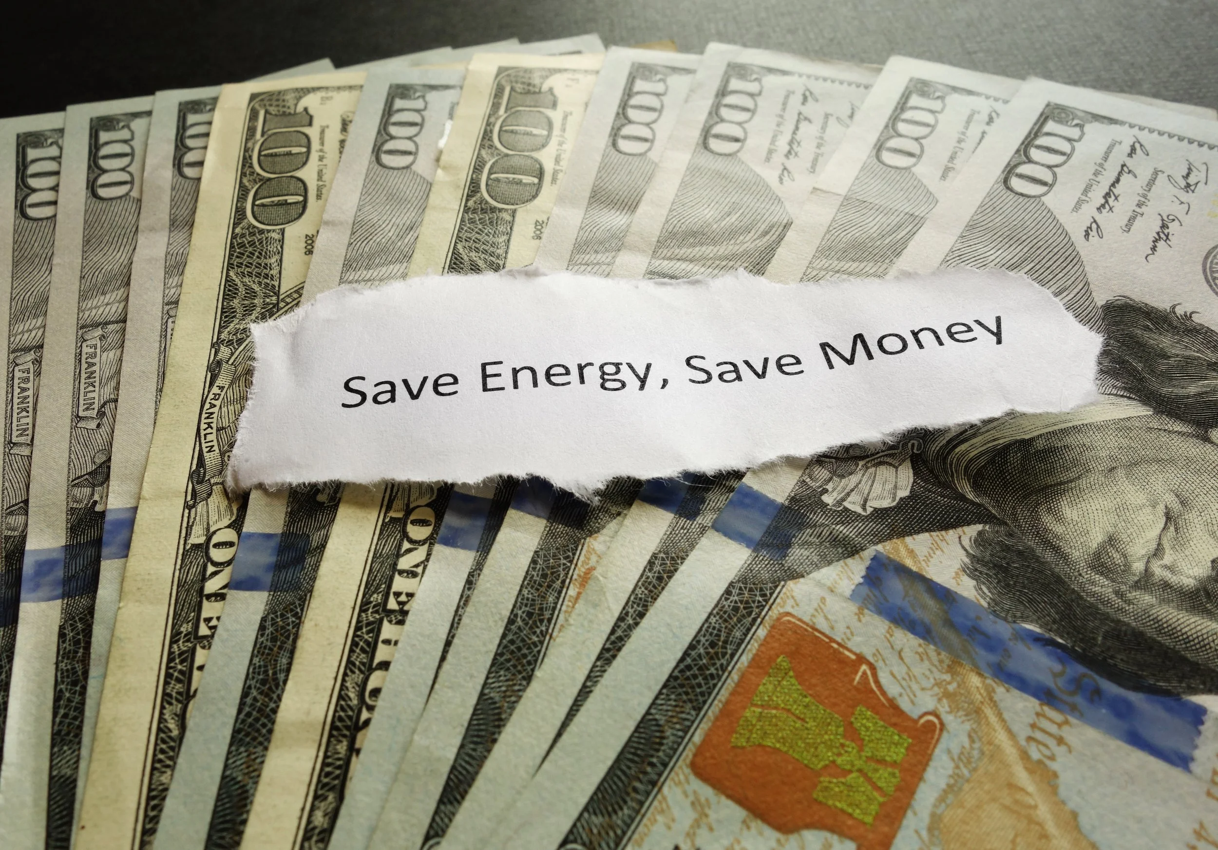 A fan of U.S. hundred-dollar bills with a torn piece of paper on top that reads 'Save Energy, Save Money'