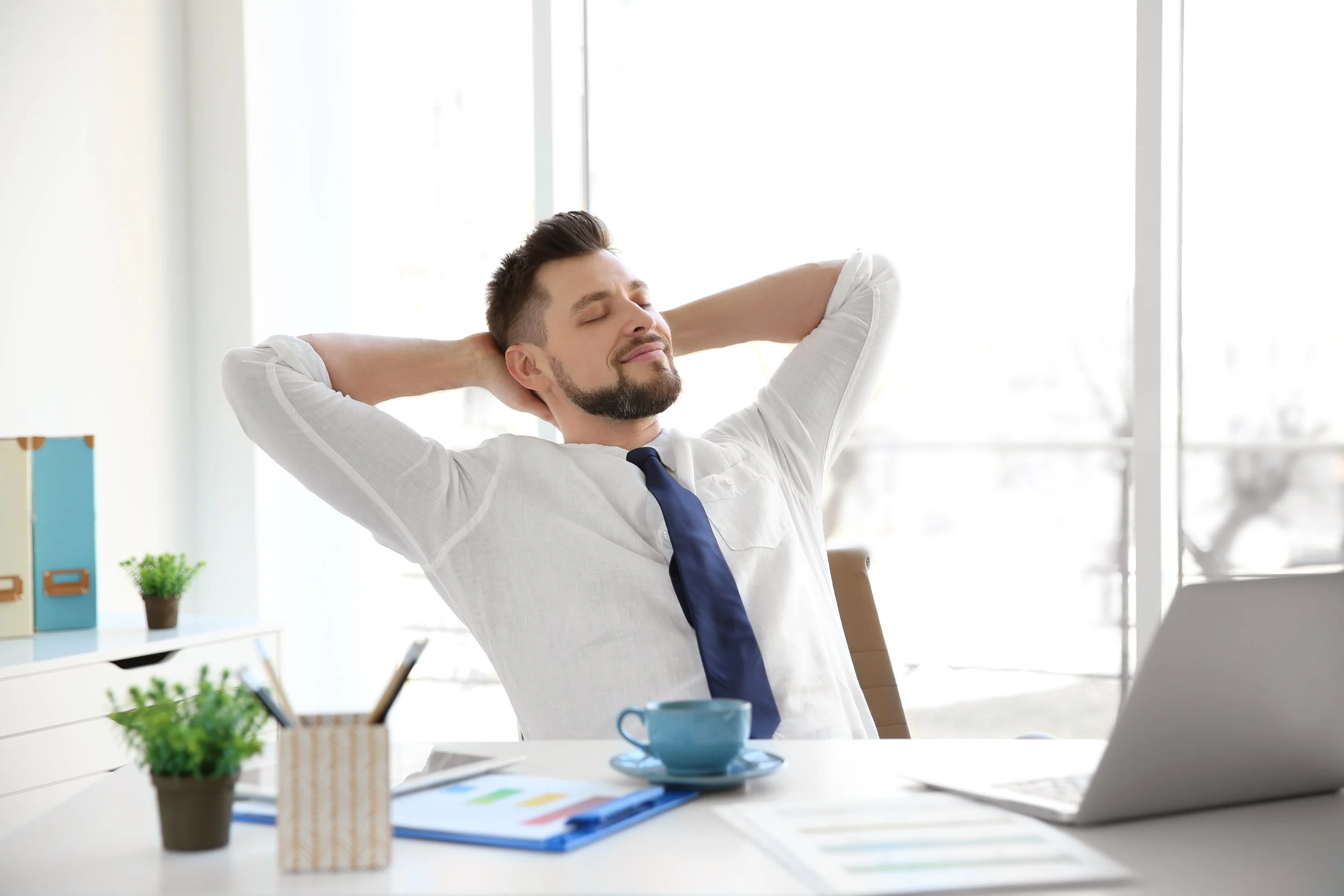 Man in office relaxing at desk with eyes closed, arms behind head, smiling, with a laptop, documents, coffee cup, and plants on desk.