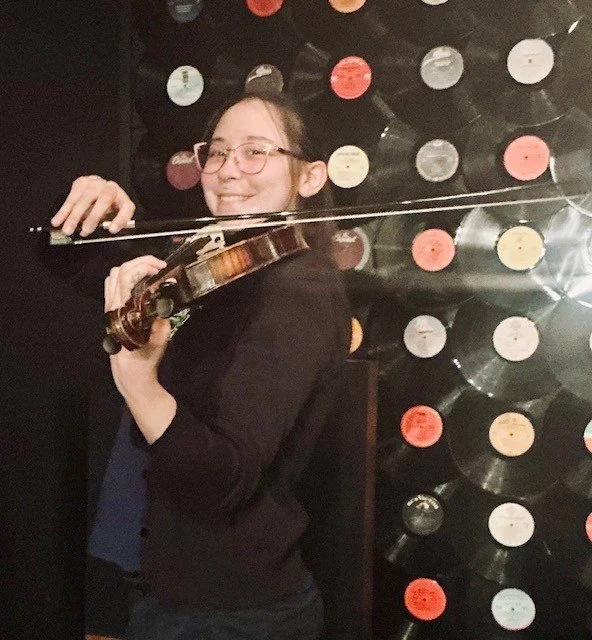 Photo of Naomi Martinez playing violin in front of a backdrop of vinyl albums.
