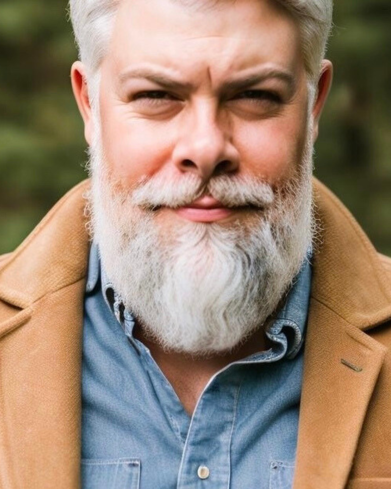 Close-up of a middle age man with white hair, gray beard, and wearing a tan coat, blue shirt, and blue jacket, with a blurred green outdoor background.