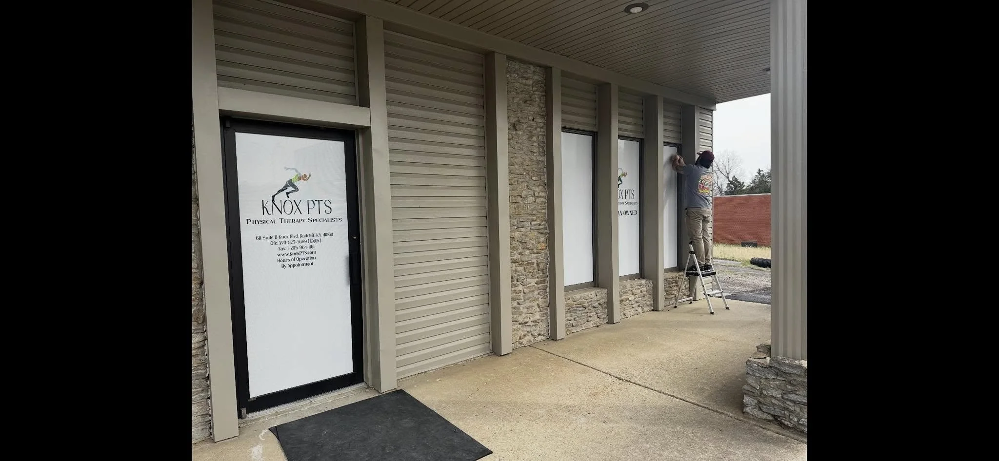 A man on a step ladder installing a window sign for Knox PTS Physical Therapy Specialists outside a commercial building with wood and stone exterior.