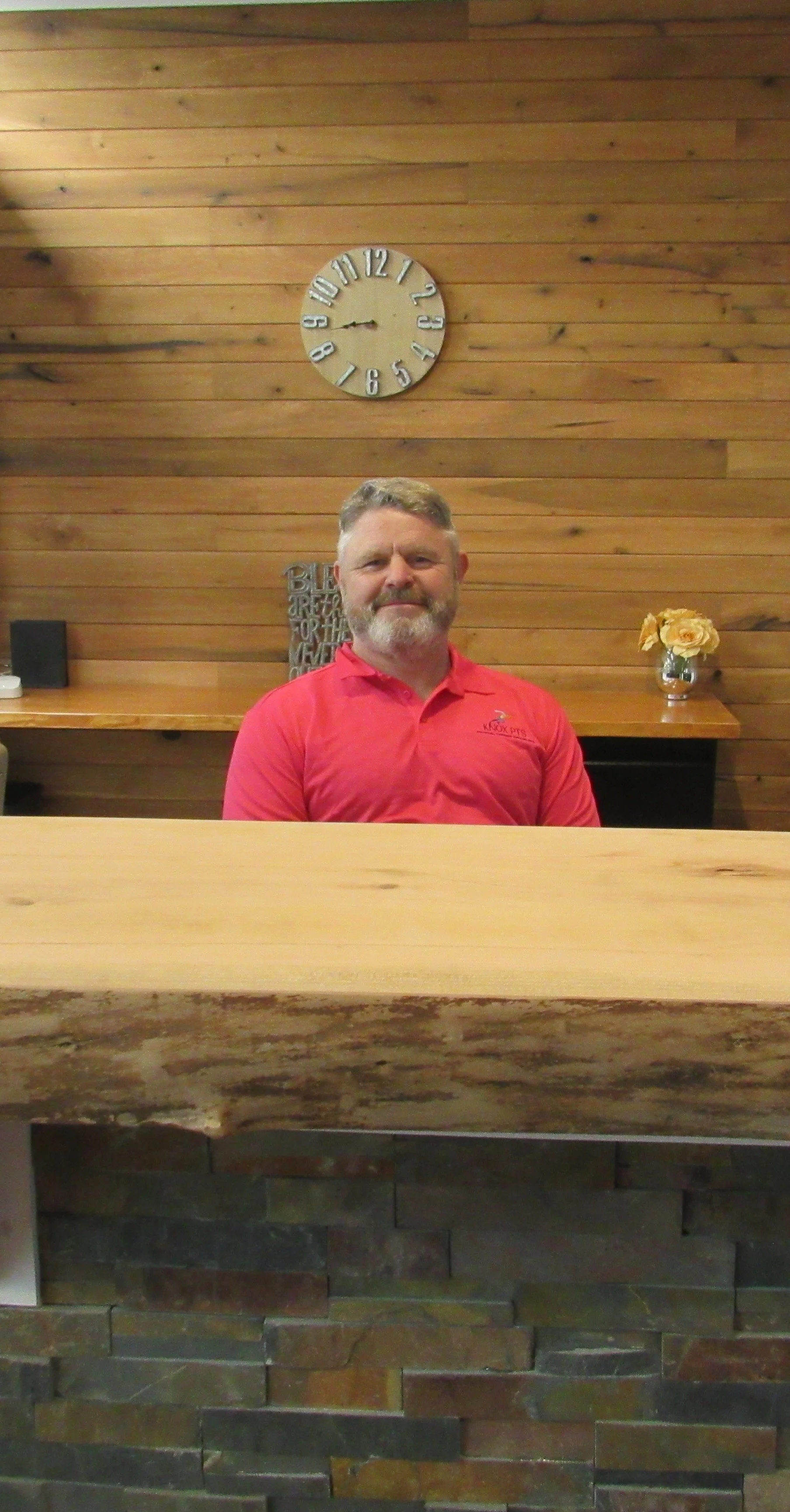 A man with gray hair and beard wearing a red polo shirt, standing behind a wooden reception desk with a stone front. The background features a wooden wall with a round clock showing 8:50, a small flower vase with yellow flowers, and a decorative sign with the word 'BLES' visible.