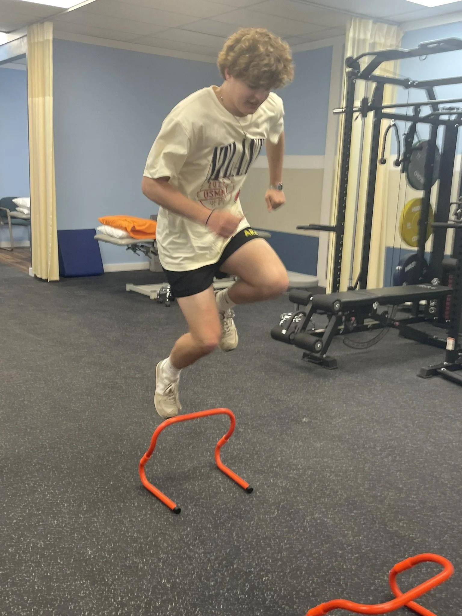 Young man with curly hair jumping over small orange hurdle in a gym. He is wearing a white t-shirt, black shorts, and white sneakers.