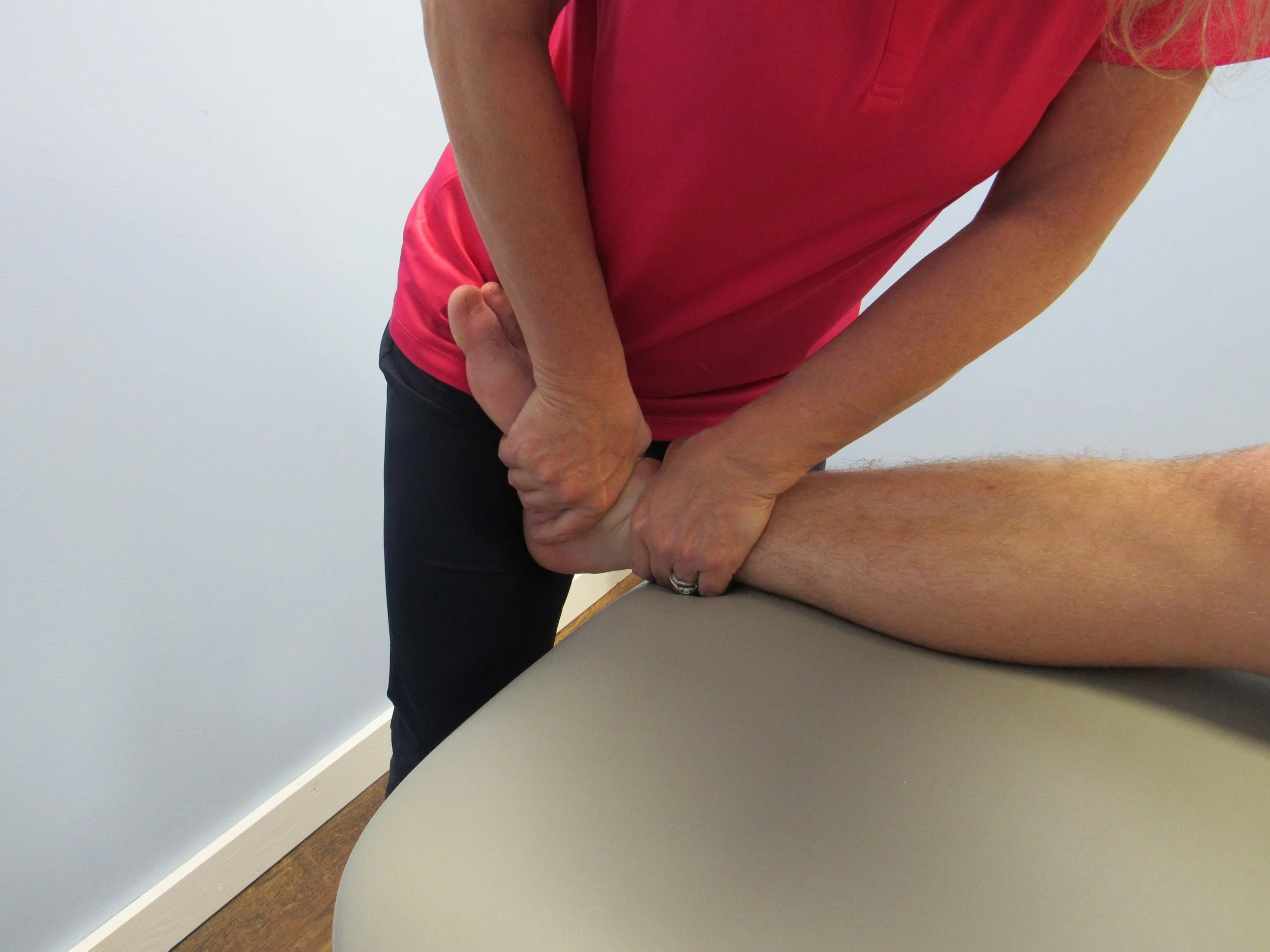 A physical therapist in a pink shirt performing a leg stretch or manipulation on a patient's leg in a clinical setting.