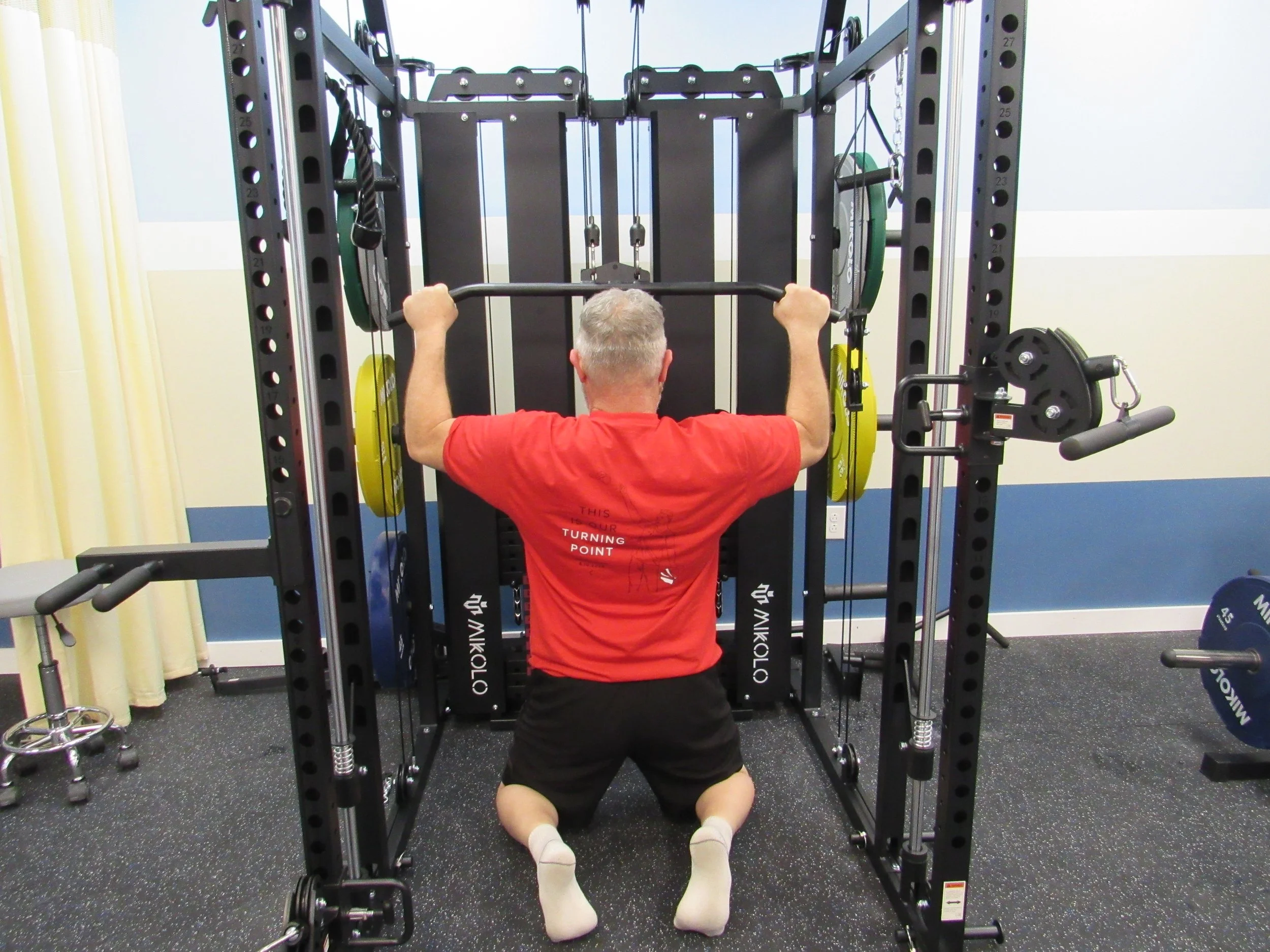 A man with gray hair kneels on the gym floor performing a shoulder press with a barbell, surrounded by a squat rack and gym equipment in a fitness center.