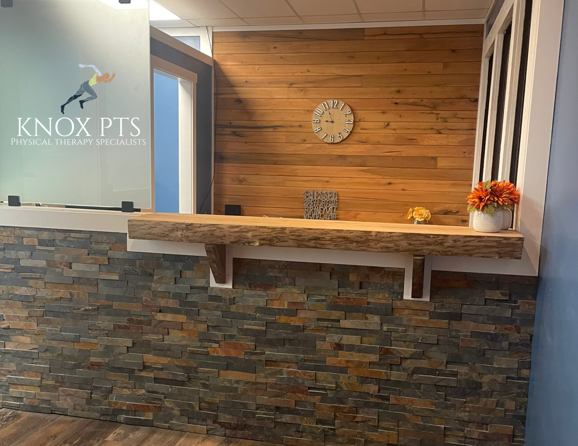 Reception area with a stone and wood counter, a wooden wall with a clock, and flower vases on the counter.