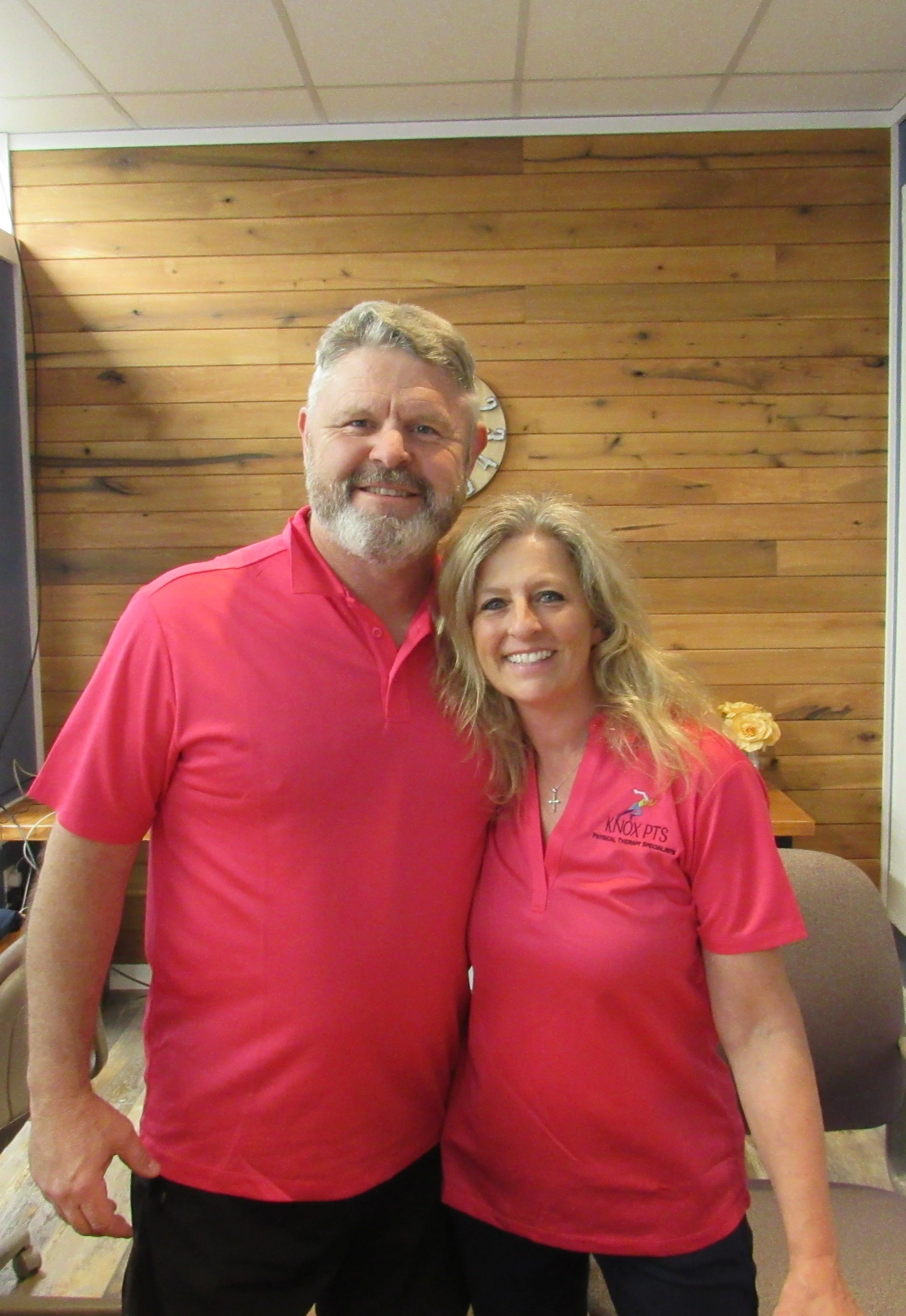 Two smiling adults, a man with gray hair and beard and a woman with shoulder-length blonde hair, standing close together inside a room with a wooden wall and clock in the background. Both are wearing red shirts.