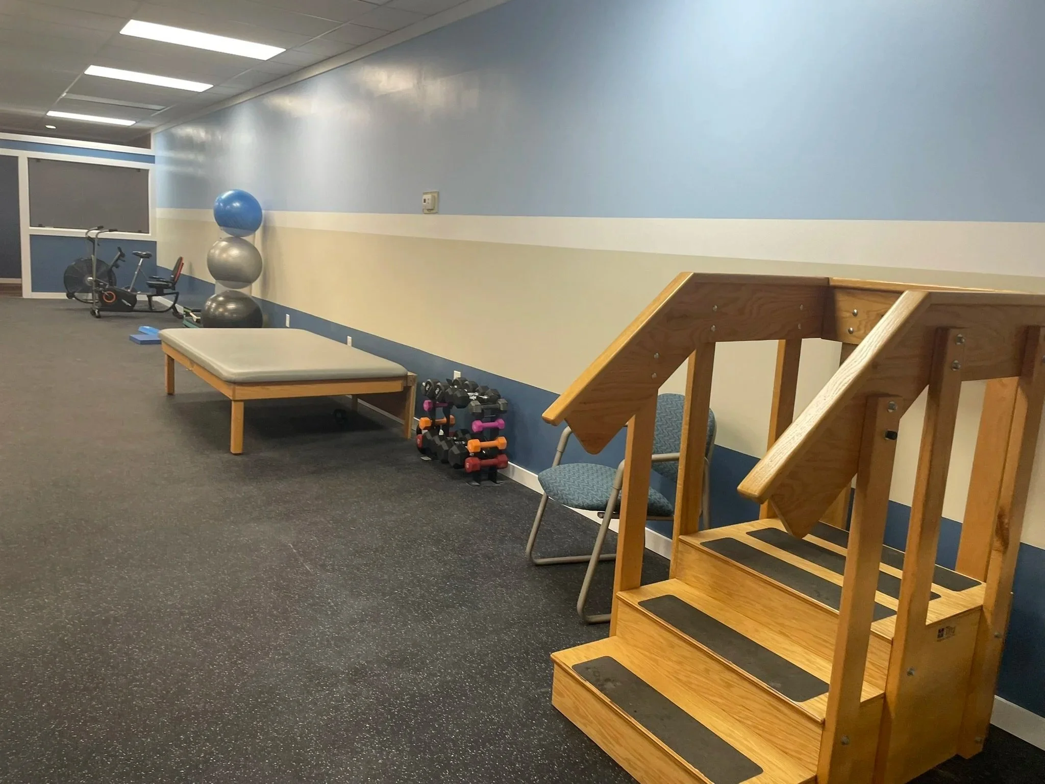 Interior of a fitness or therapy room with exercise equipment, including exercise balls, dumbbells, and a step platform, and a wooden stair climber on a dark speckled floor.