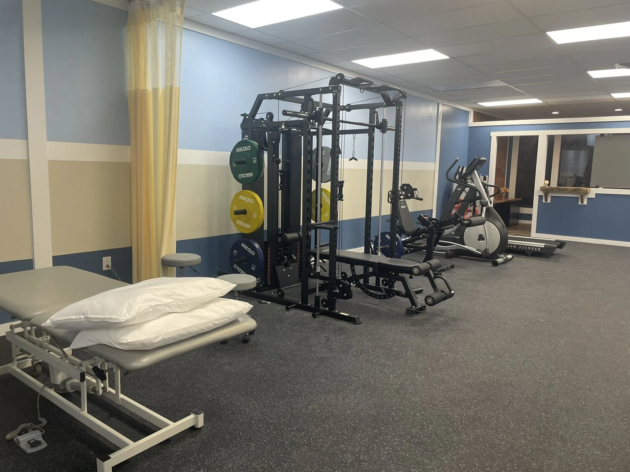 A room with medical and fitness equipment, including an adjustable examination table with pillows, a weightlifting station with colorful weights, an exercise bike, and a treadmill, with blue and yellow walls and yellow curtains.