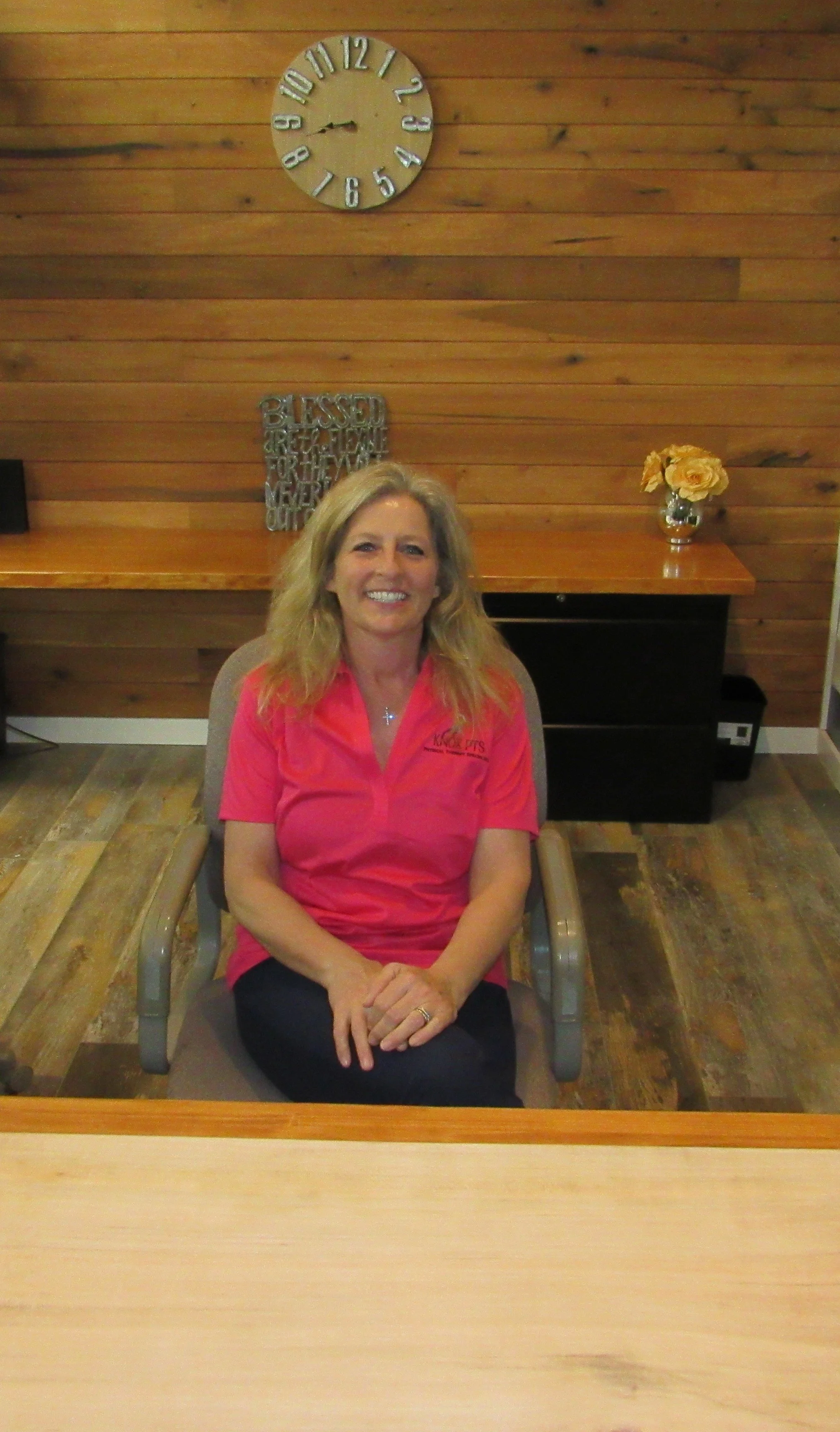 A woman with blonde hair, wearing a pink shirt with a logo, sitting in an office chair in front of a wooden wall with a clock, a decorative sign, and a vase with flowers on a desk behind her.