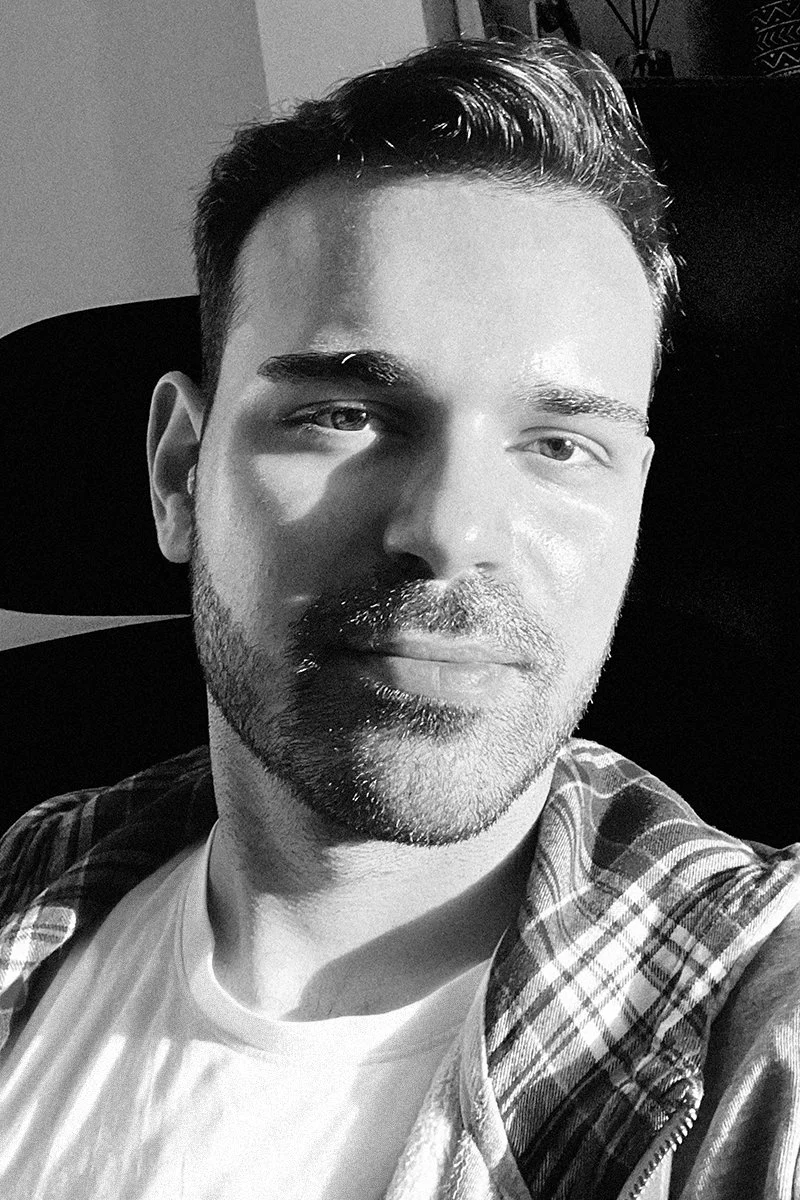 A black-and-white photo of a young man with dark hair and a beard, sitting against a dark background, wearing a checkered jacket over a white shirt, looking at the camera with a slight smile.