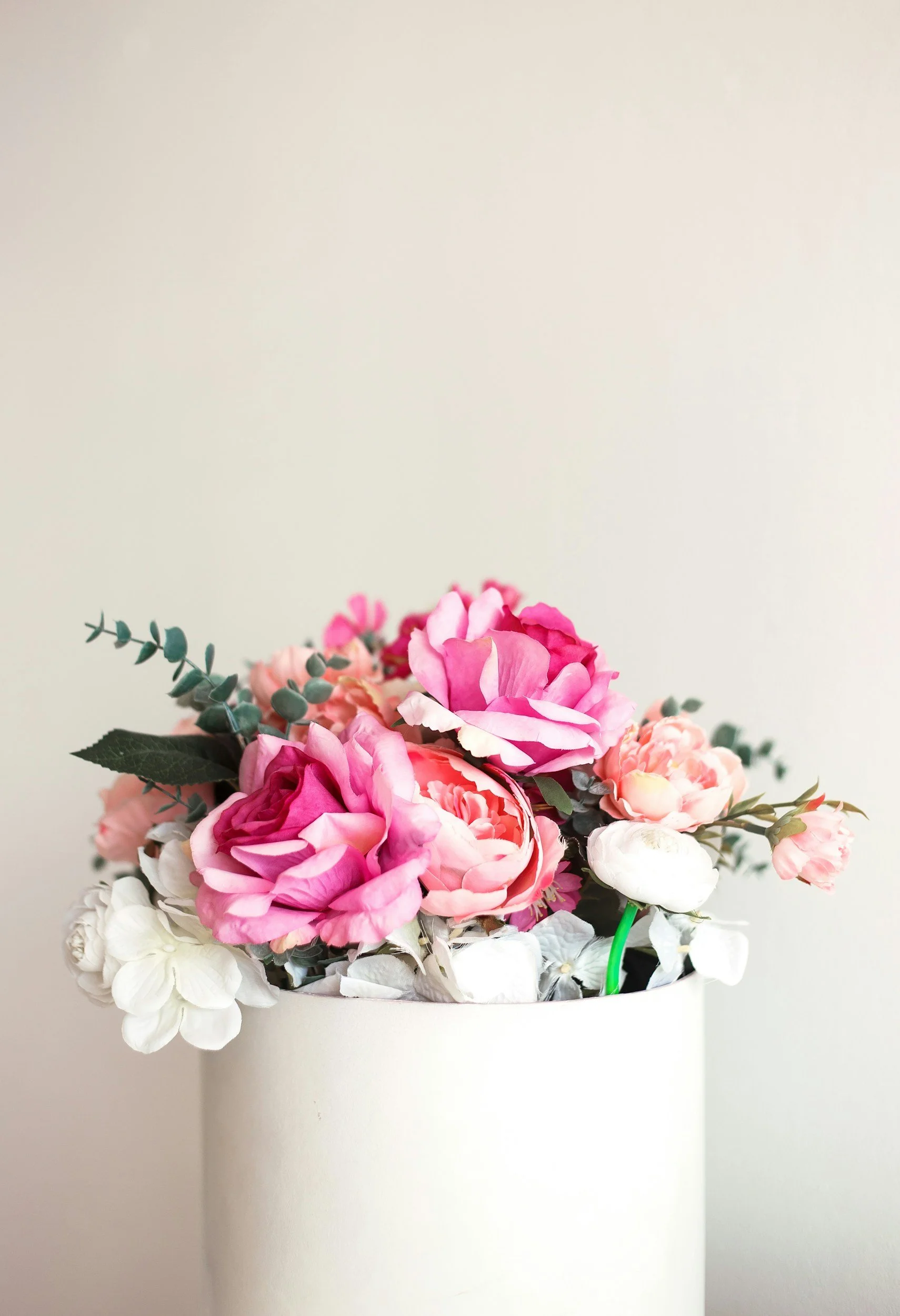 A white vase with pink and white artificial flowers, including peonies and eucalypt leaves, against a plain wall.