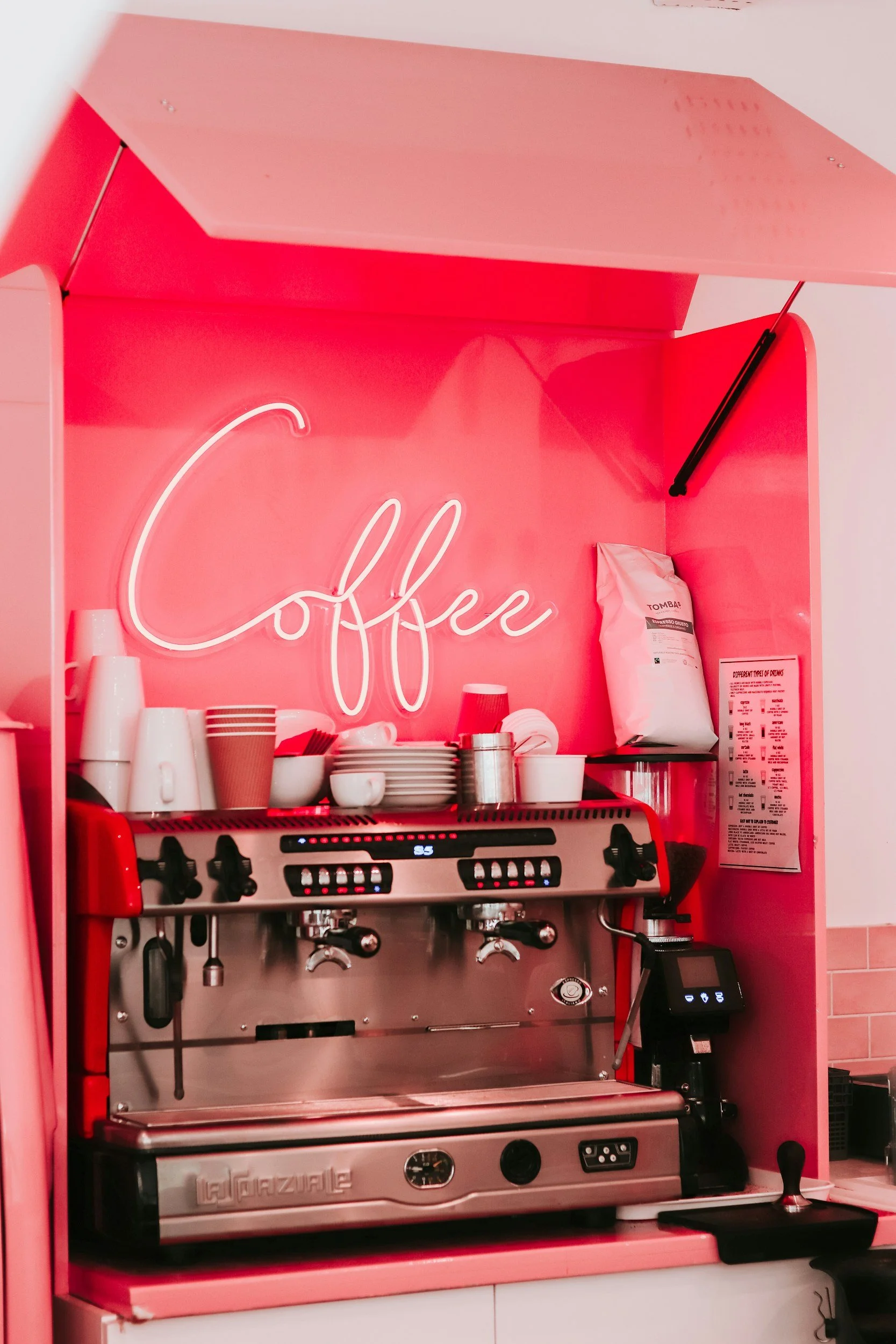 Pink coffee station with a neon sign that says 'Coffee', an espresso machine, cups, cups on a shelf, and a bag of coffee beans.
