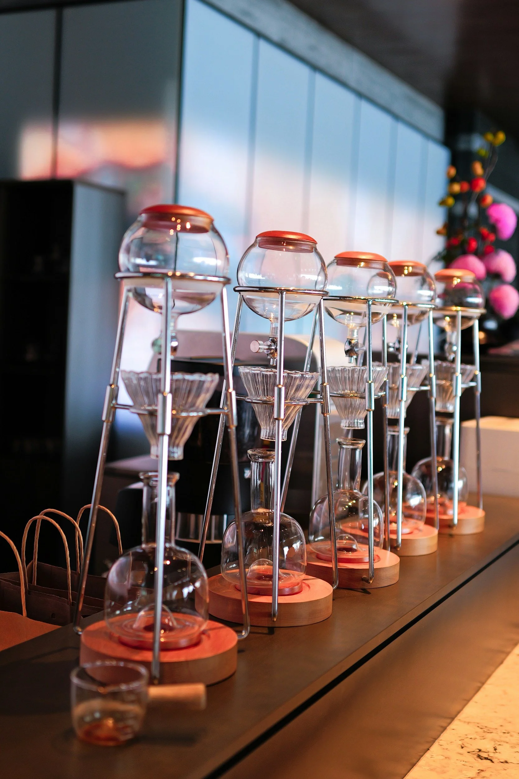 A row of glass siphon coffee makers on a dark countertop in a modern setting with a colorful, decorated wall in the background.