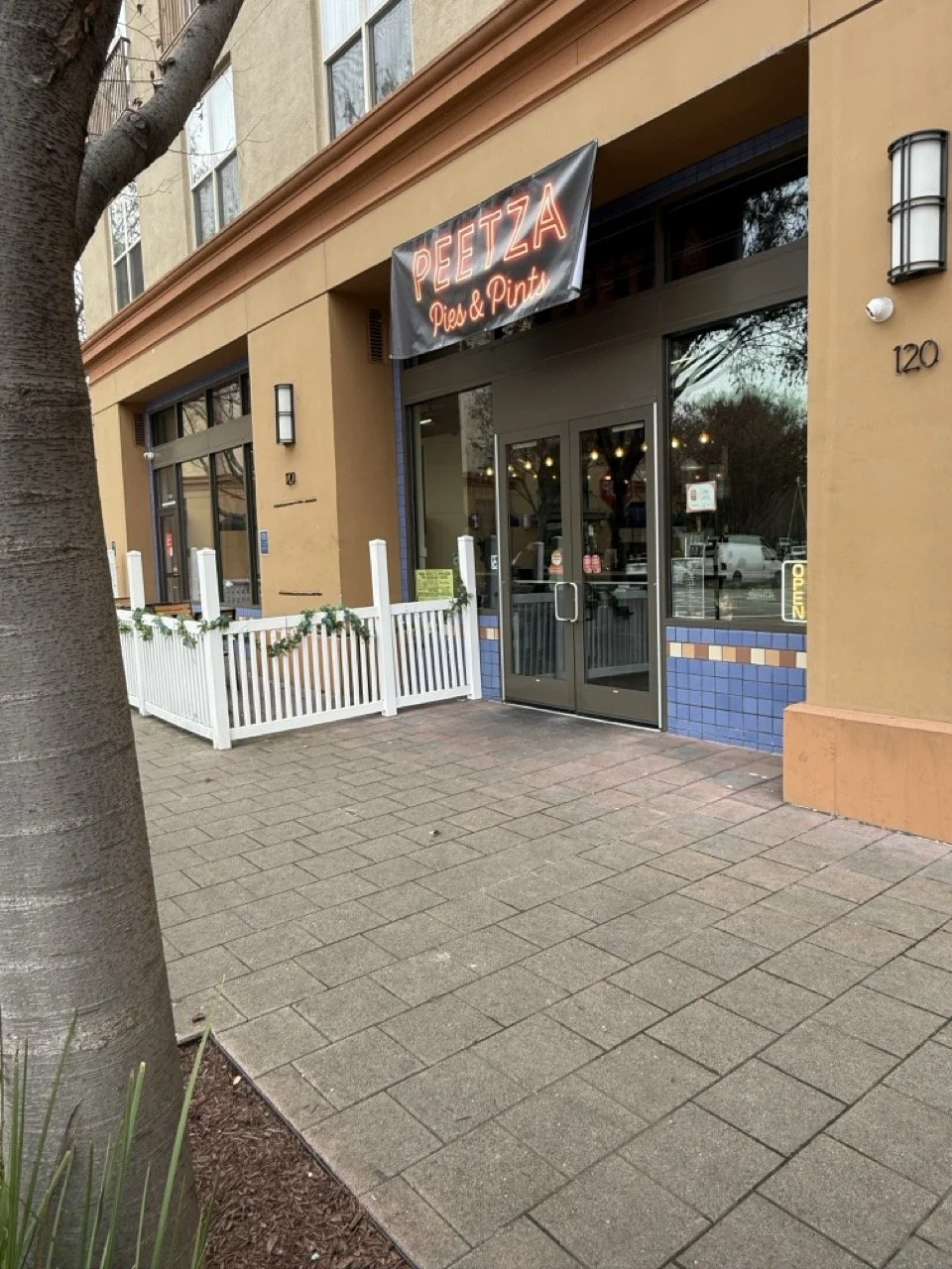 Exterior view of a restaurant named Peetza Pie & Pint with a black sign displaying the name in red neon lights. The entrance has glass double doors, and there is a white picket fence decorated with greenery outside. The building has a beige facade with blue tile accents near the door, and the street number 120 is visible on the wall. There is a tree on the sidewalk to the left.
