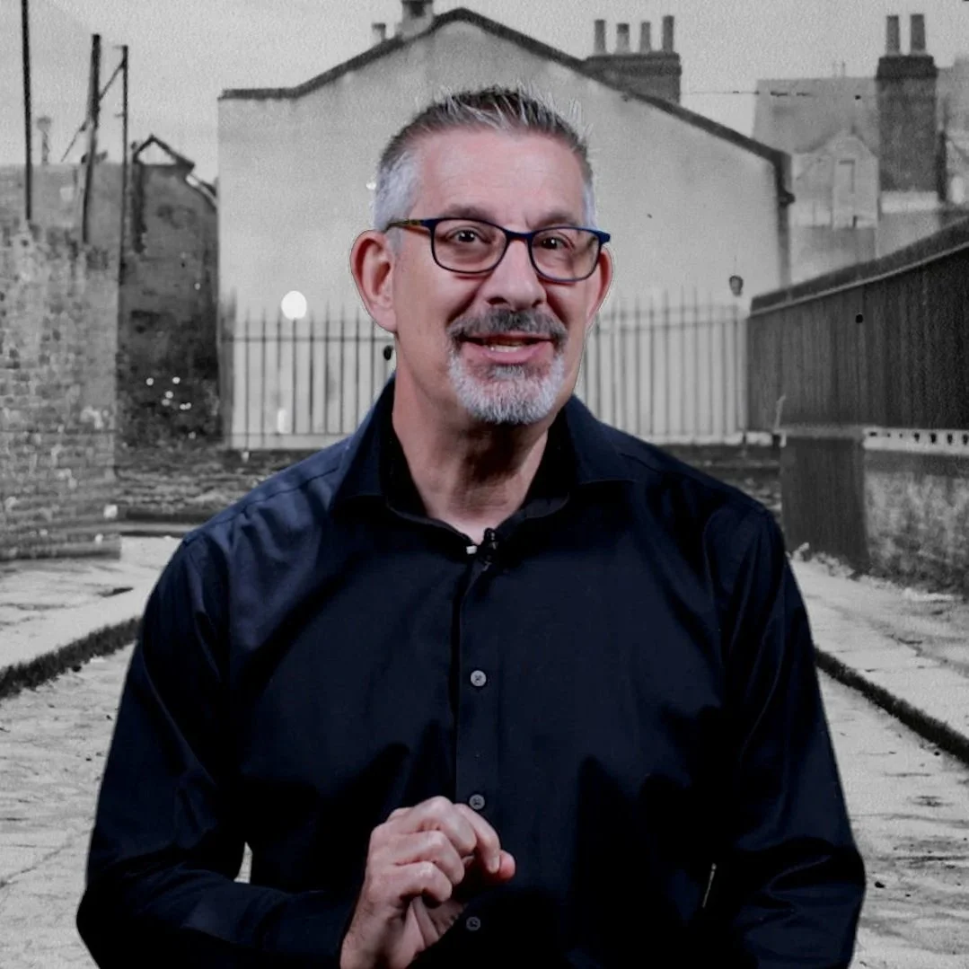 A man with glasses, gray hair, and a goatee smiling and holding his jacket lapel, standing in front of a black and white photo of a city alleyway with buildings and fences.