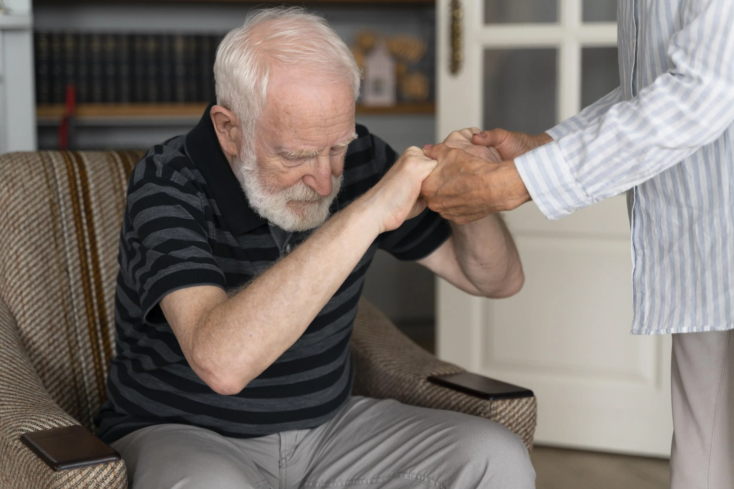 An elderly man with white hair and beard holding hands with a caregiver in a bright room, one person sitting on a patterned armchair.
