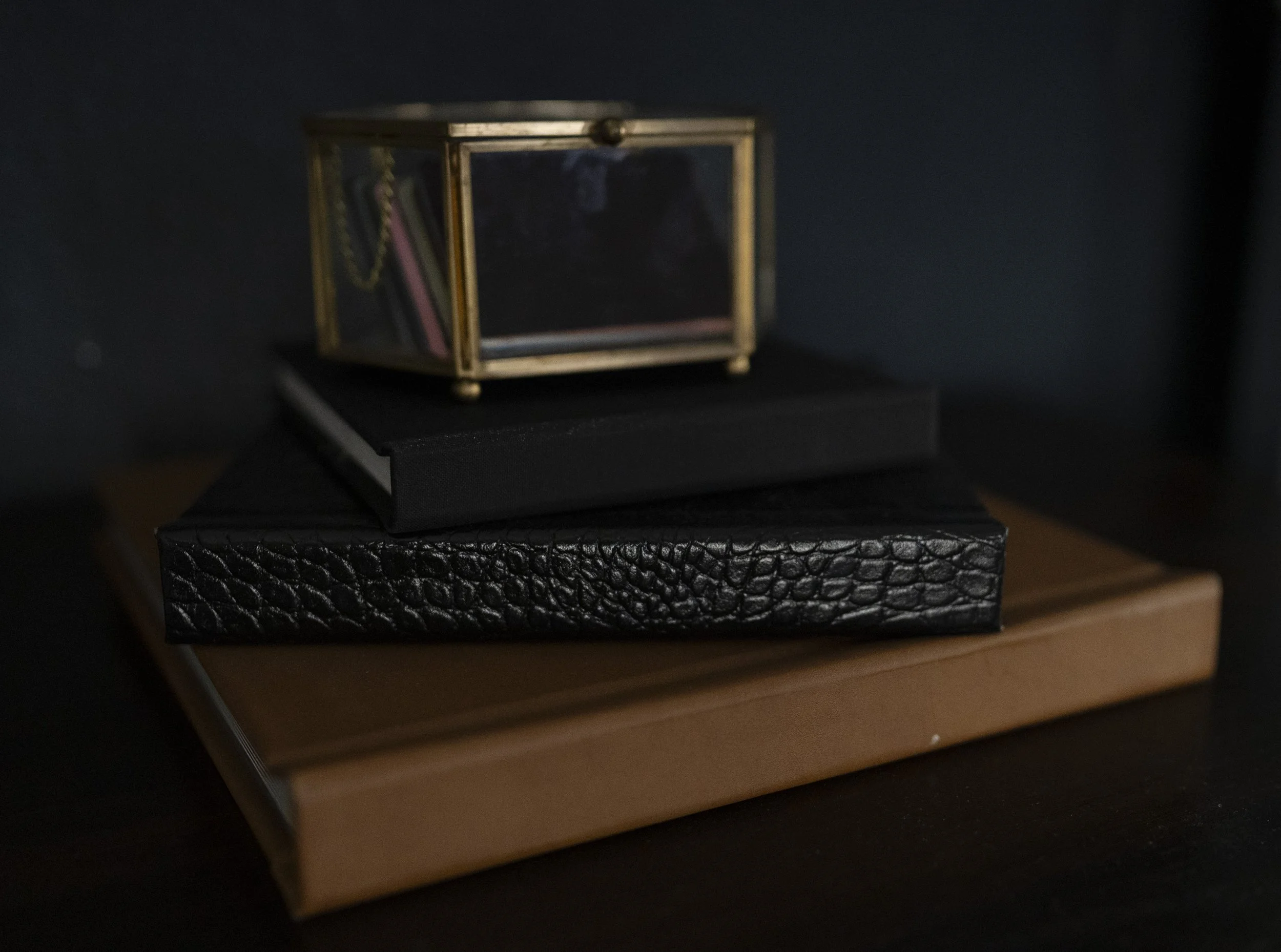 Stack of three books with a small gold framed glass box on top, containing jewelry, placed on a dark surface.
