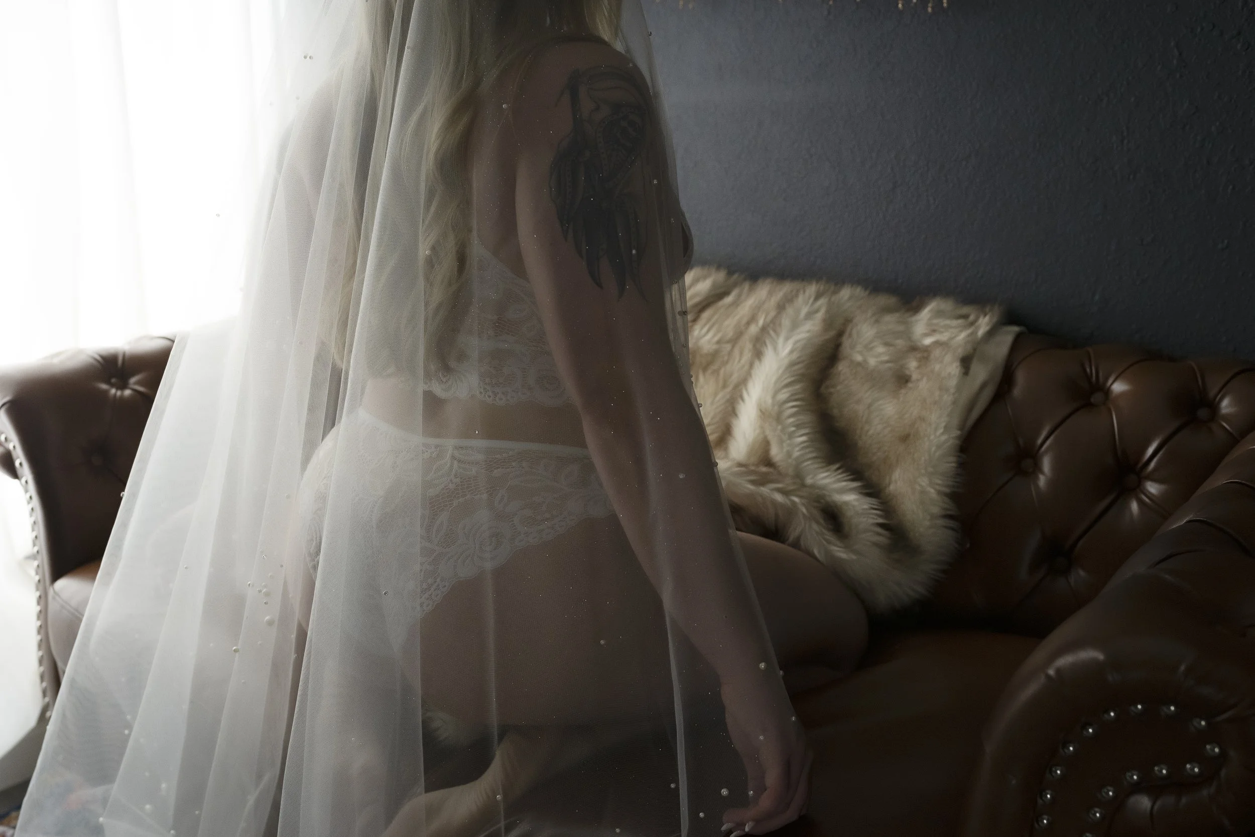 A woman in lace lingerie with a veil sitting on a leather couch, with a fur throw and fur pillow in the background.