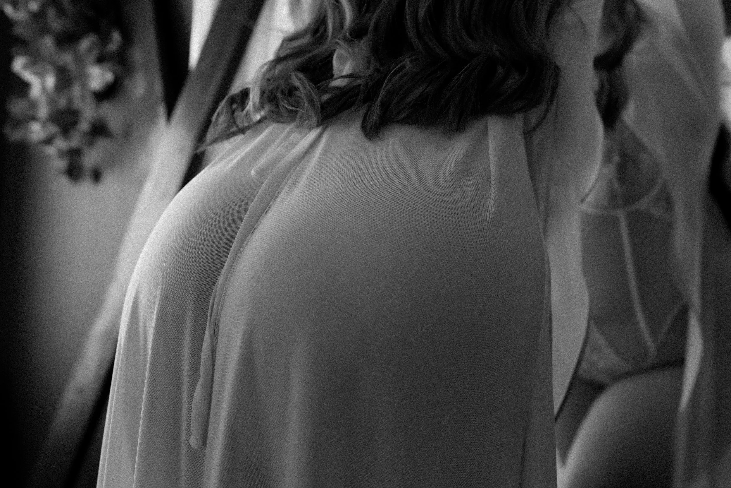 Close-up of a woman with dark, wavy hair wearing a light-colored sleeveless dress, seen from behind.