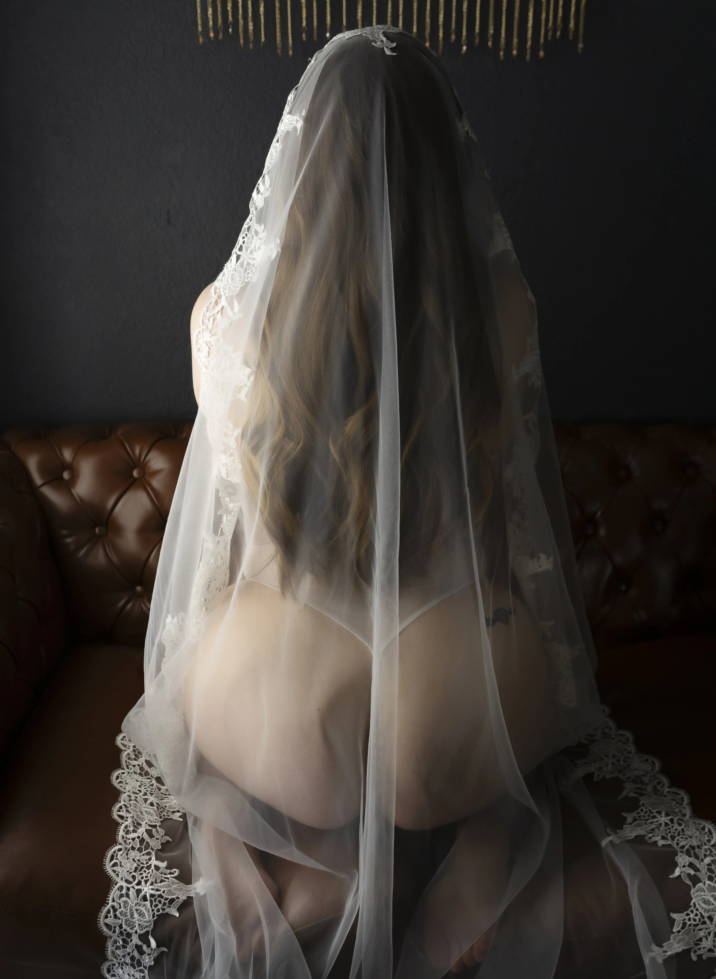 A woman with long brown hair, dressed in lingerie, seated on a brown leather couch, covered with a sheer veil with lace details, facing away from the camera.