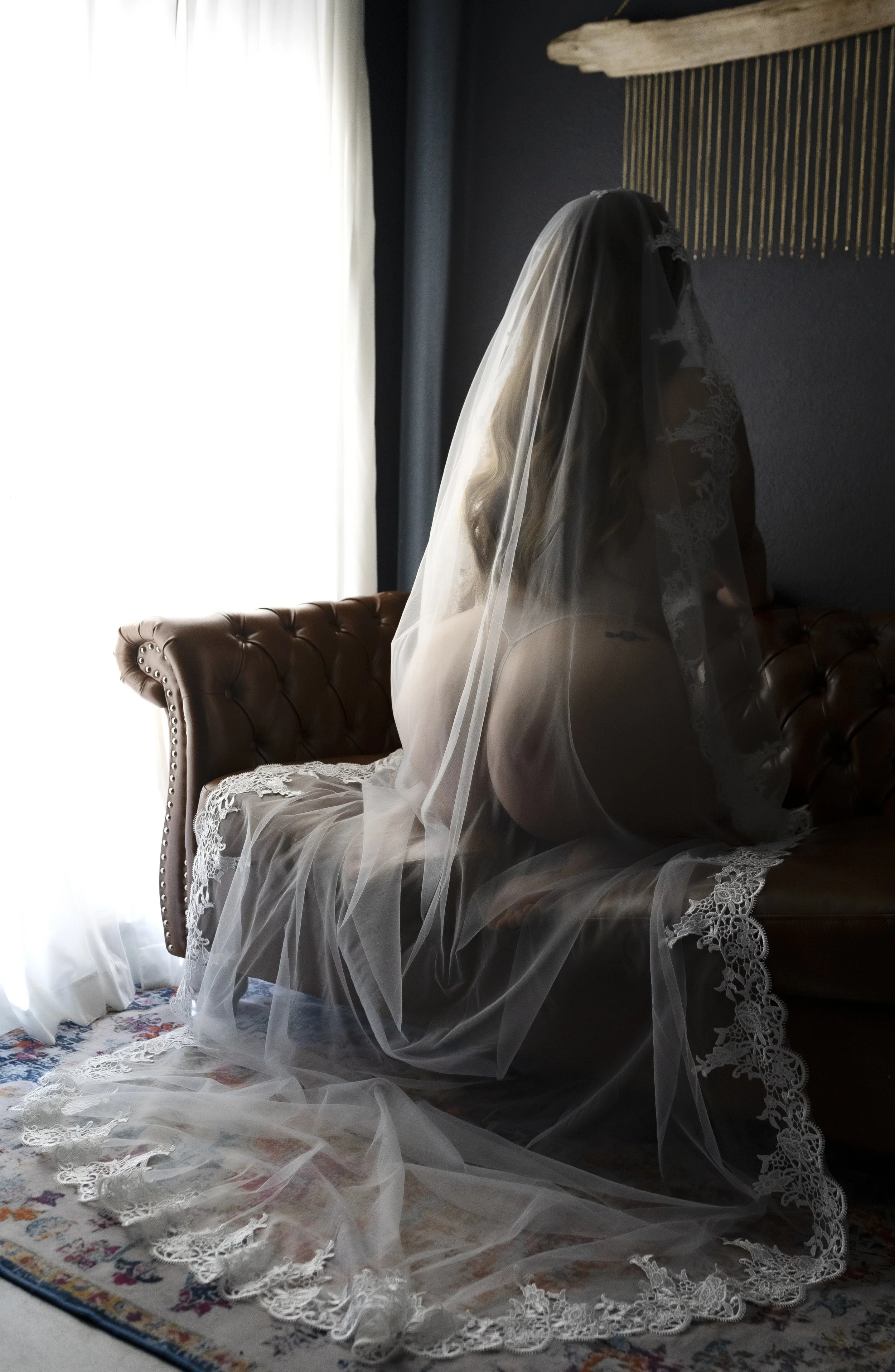 A woman sitting on a vintage leather sofa, dressed in lingerie with a lace veil over her face, in a softly lit room.