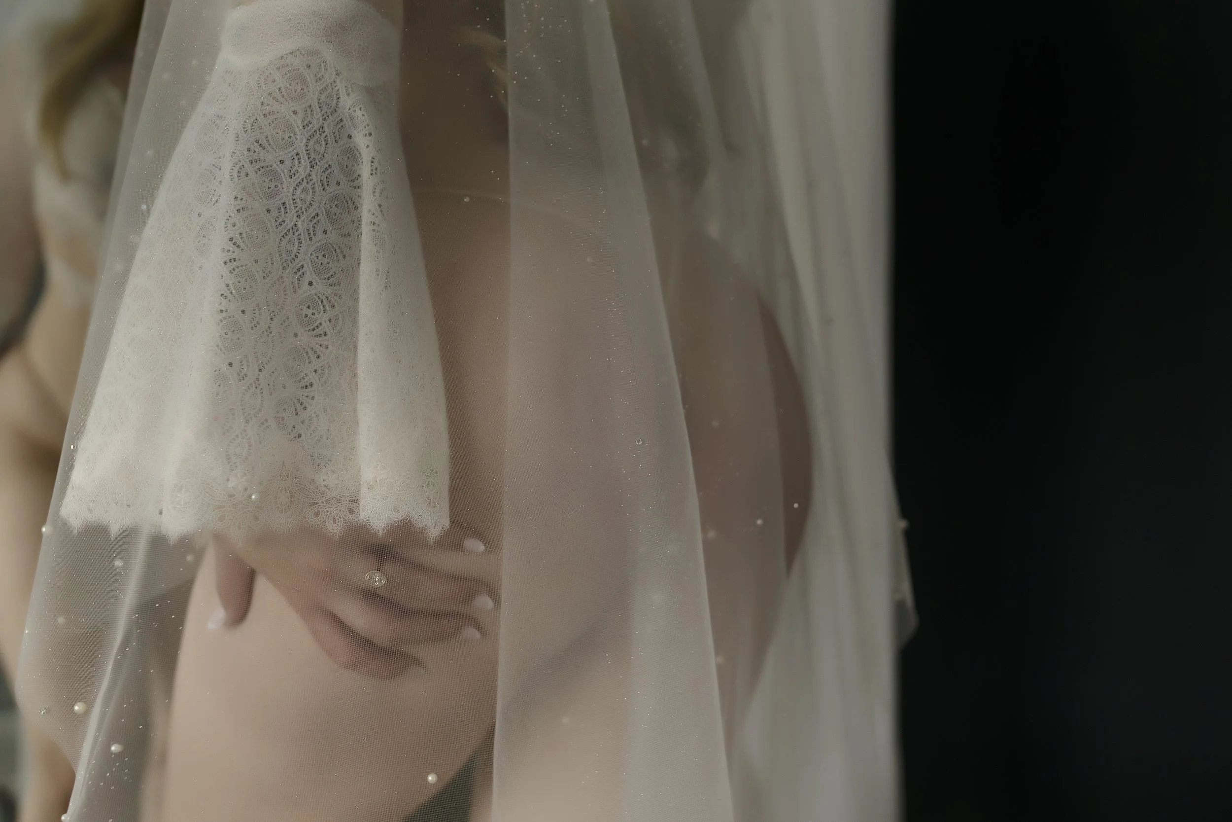 A bride wearing a lace wedding dress and veil holding her pregnant belly, visible through the sheer veil with a black background.