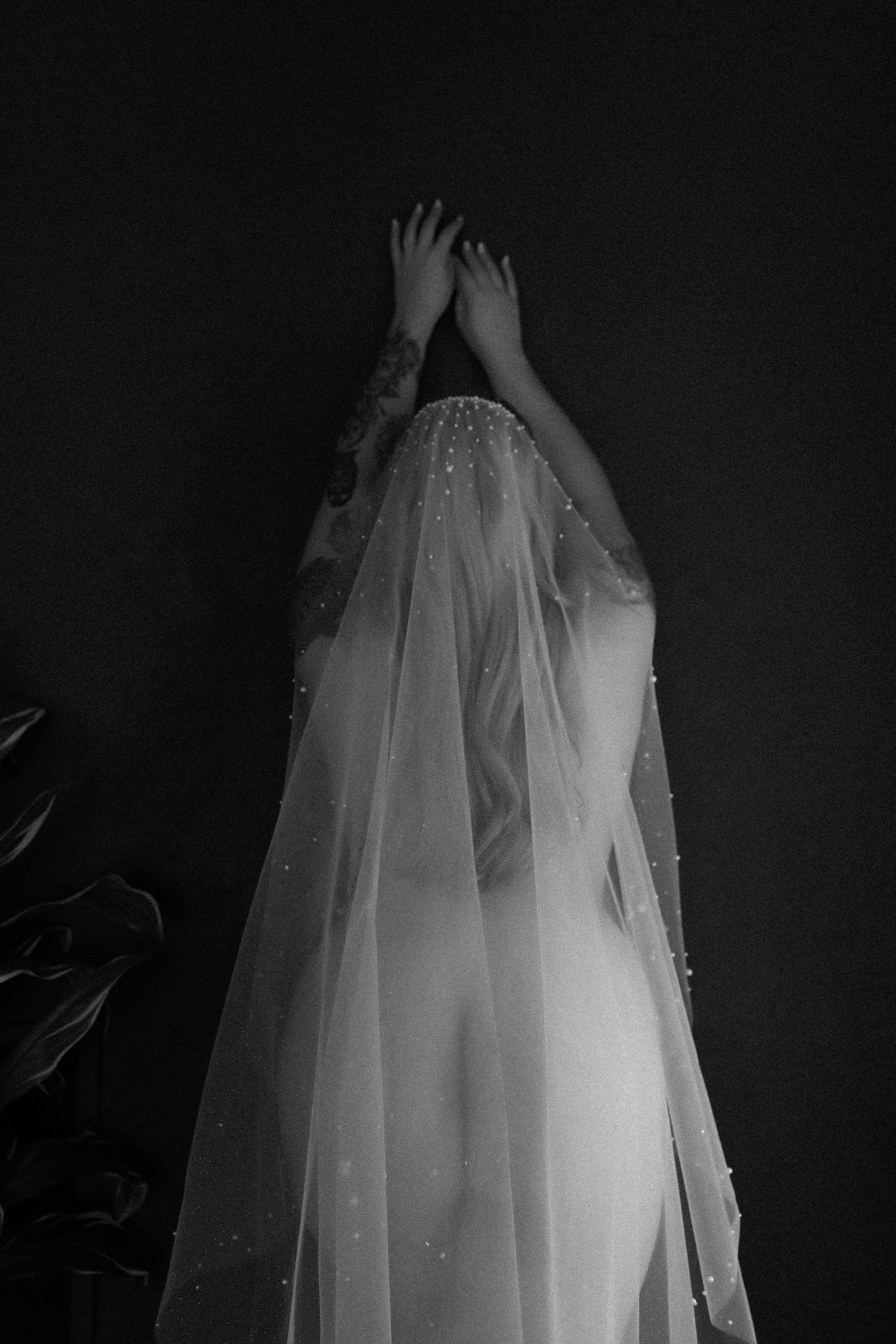 A bride dressed in a wedding gown with a veil, her hands covering her face, facing a dark background.