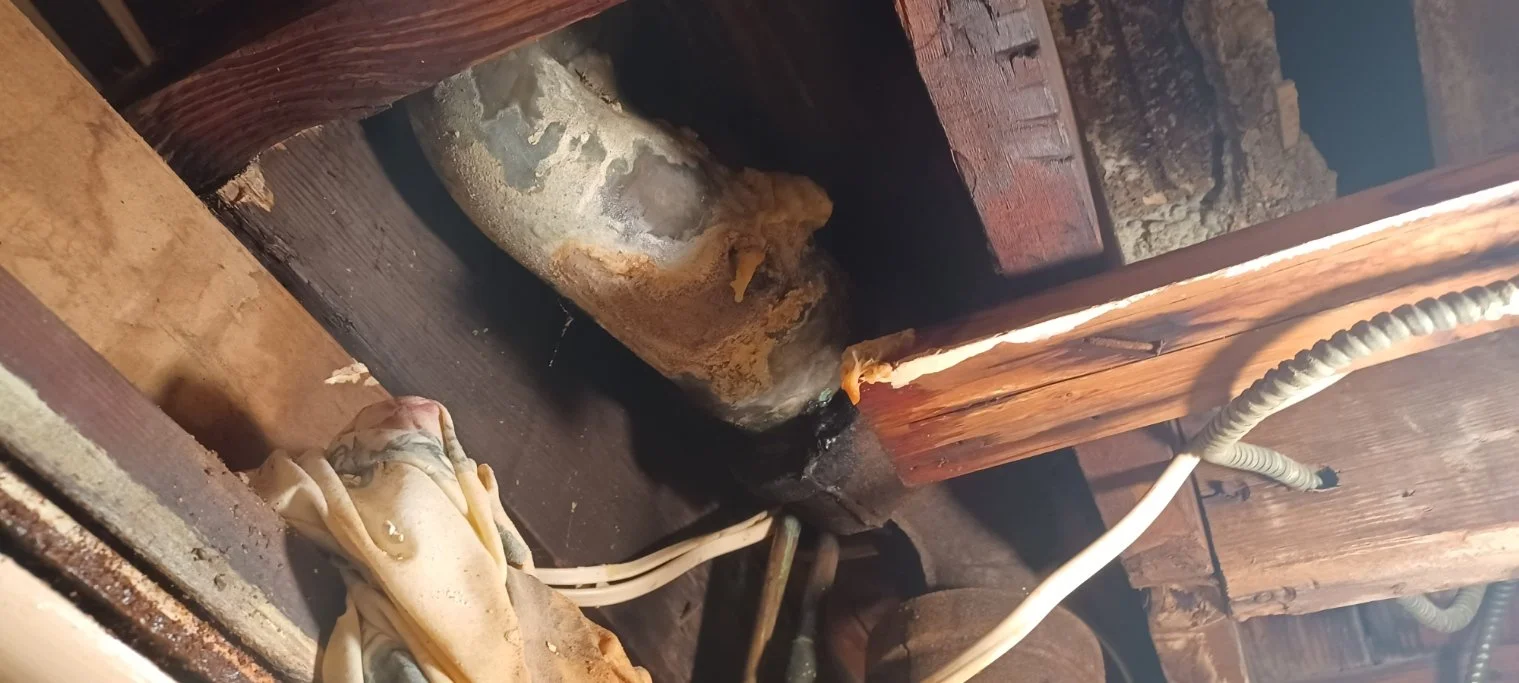 Close-up of a wood-framed space showing rusted metal pipe, electrical wiring, and corroded wood.