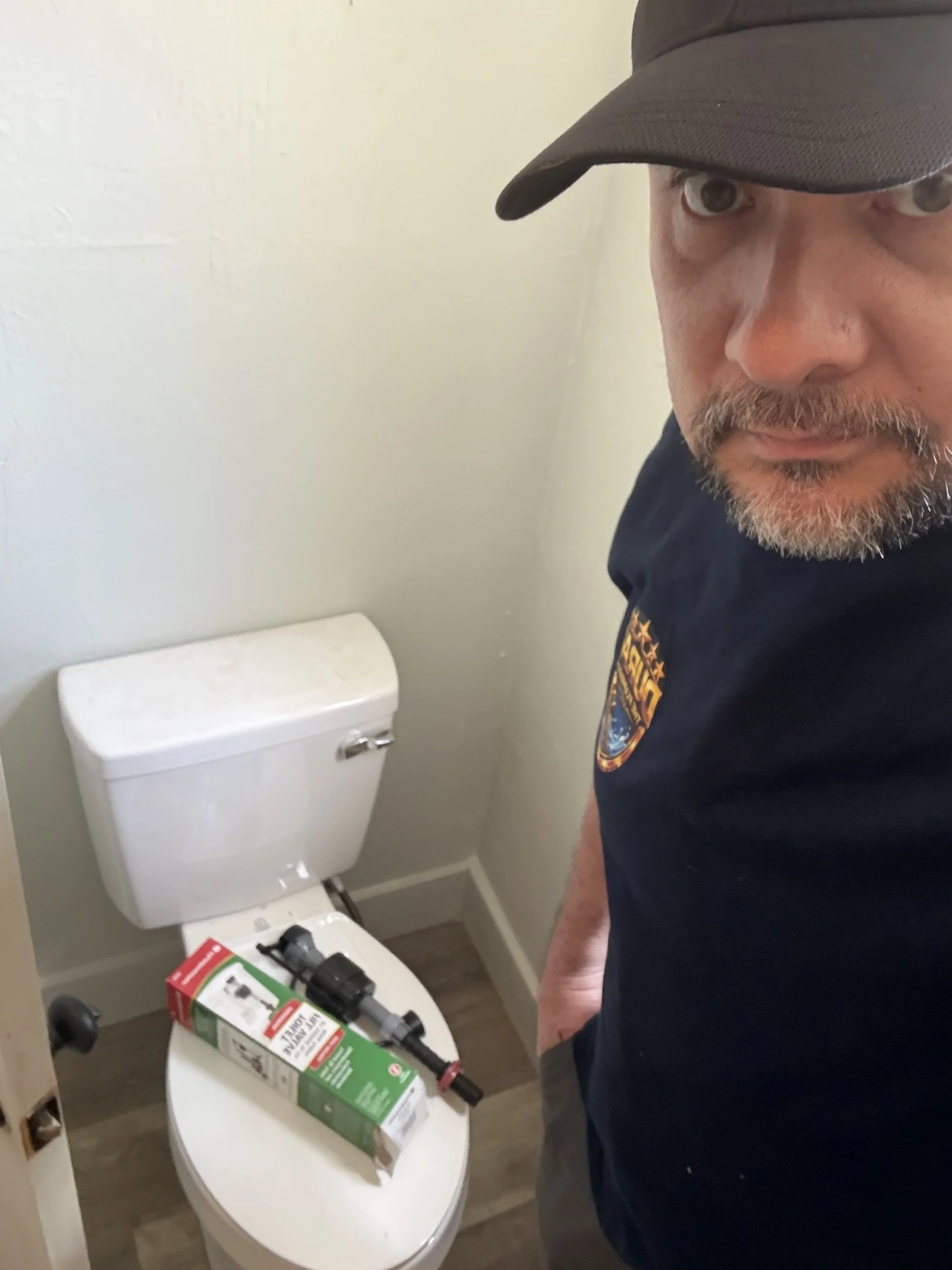 A man wearing a black cap and black shirt with a patch on his sleeve stands next to a white toilet with various items on the tank, including a box, a black flashlight, and black gloves. The man's face is partially visible, showing his eyes and beard.
