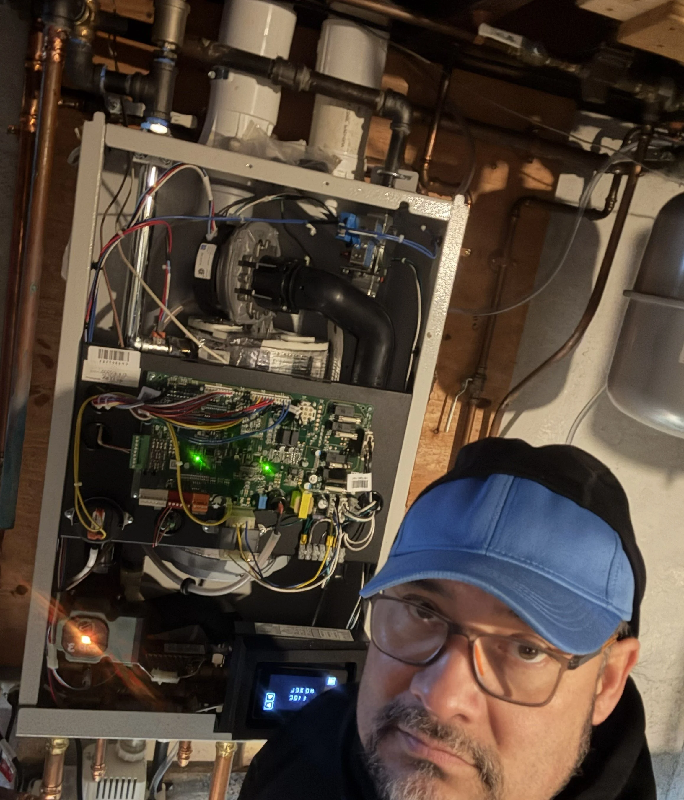 A man with glasses and facial hair leaning over a furnace or boiler system with visible pipes, wires, and electronic components inside an industrial or utility room.