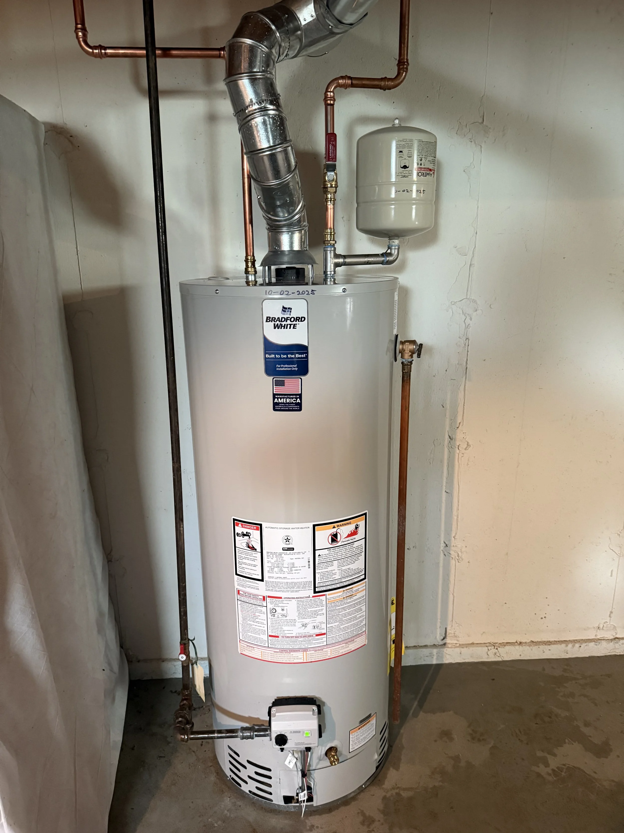 A standing white Bradford White water heater with a metal exhaust pipe, copper pipes, and a small expansion tank mounted on the wall.