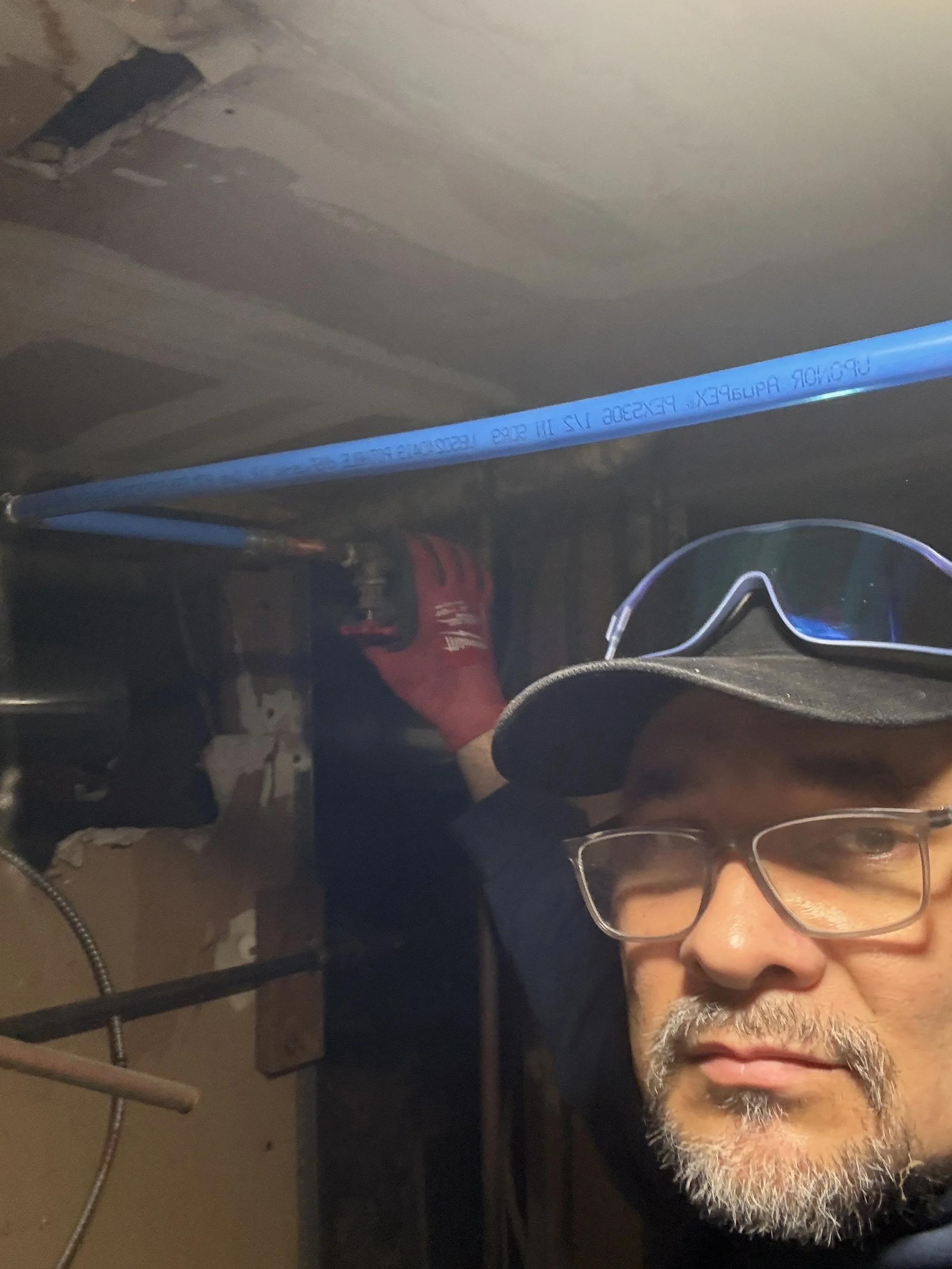 A man wearing glasses, a cap, and red gloves working on pipes in a basement or crawl space.