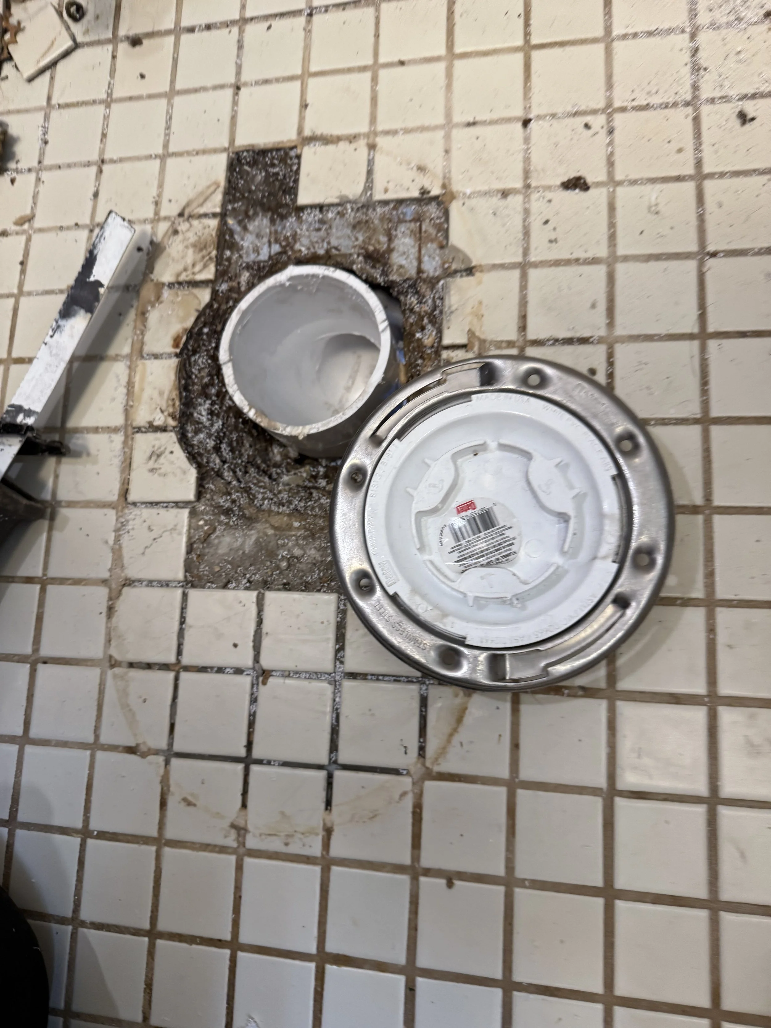 Disassembled toilet flange in a bathroom with removed toilet, revealing the drain pipe and drain flange on a tiled floor.