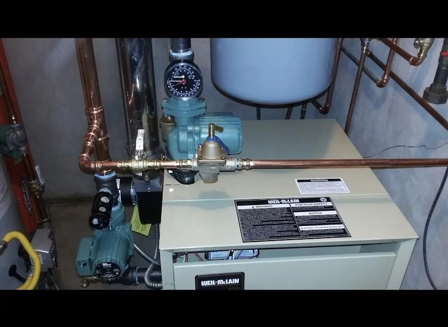 A boiler system with copper pipes, a pressure gauge, a teal pump, and a large white tank in a basement or utility room.