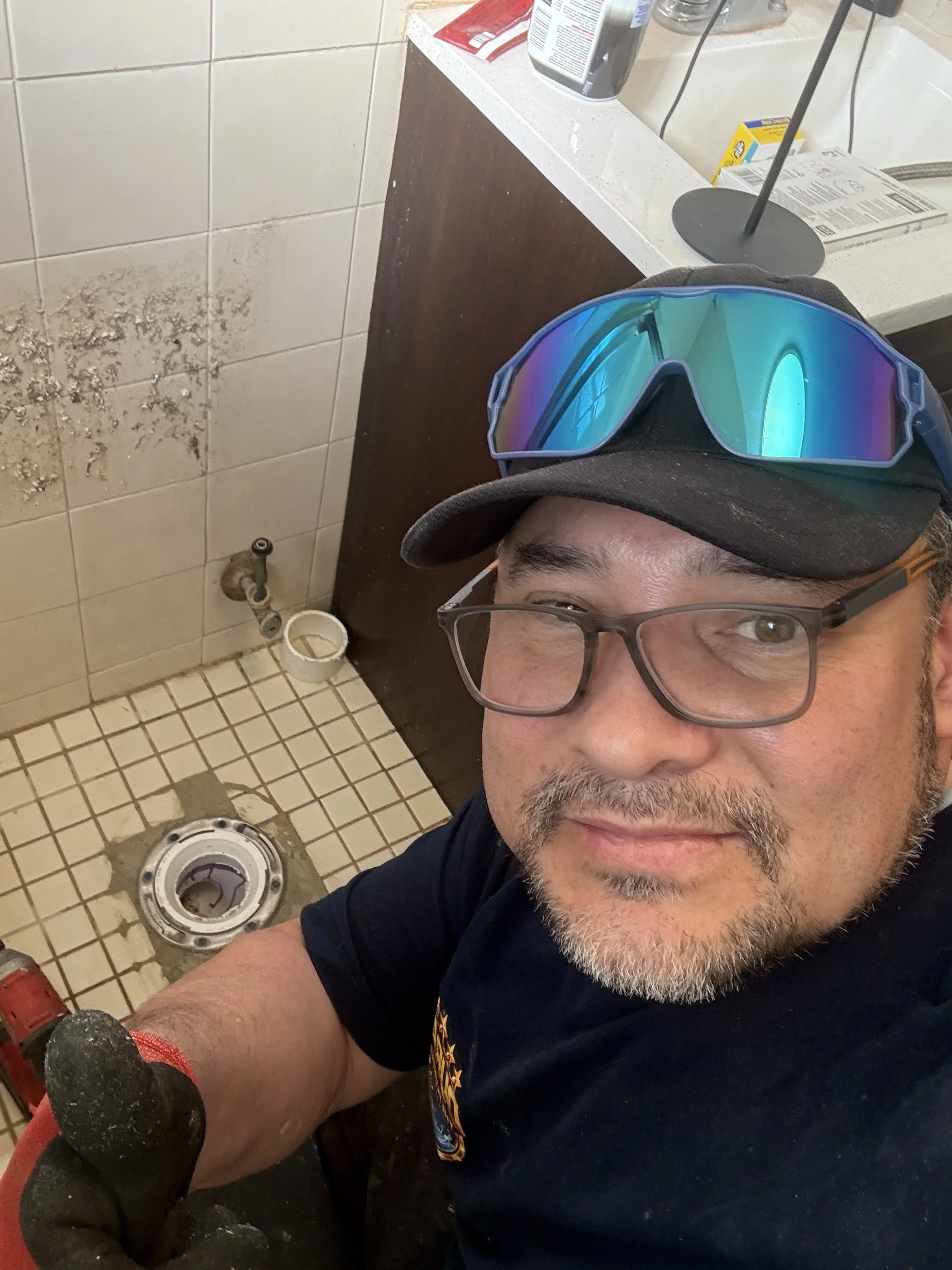 Man taking a selfie in a bathroom, wearing glasses, a black cap with reflective sunglasses on top, and gloves, while repairing or installing a toilet.