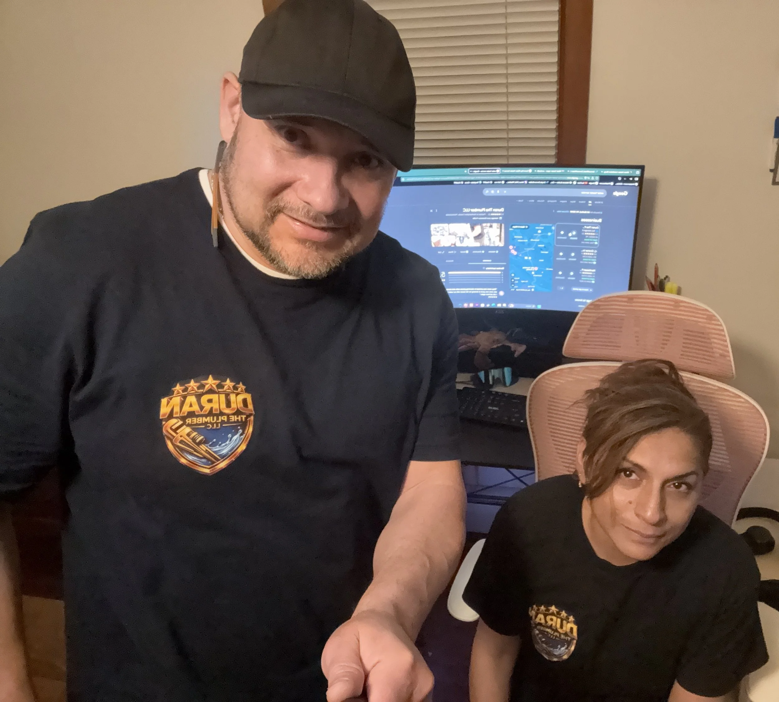 A man and woman in matching black T-shirts with a logo, posing in a home office with a computer monitor in the background.