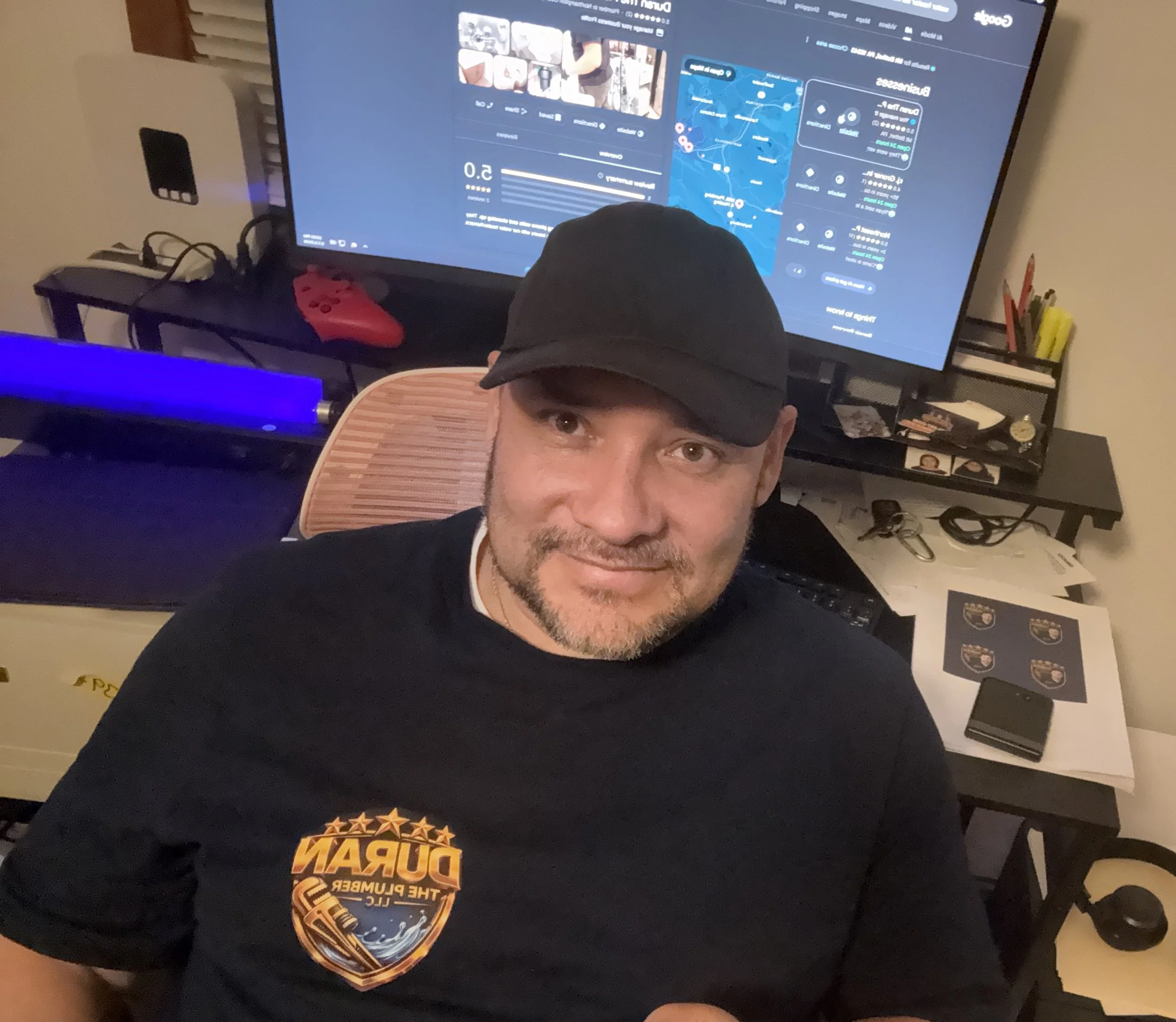A man wearing a black cap and BURN shirt sitting in front of a computer desk with a large monitor displaying a map and data. The desk has various items including a pair of headphones, a phone, and documents. The background shows a black shelf with office supplies and a red gaming controller.