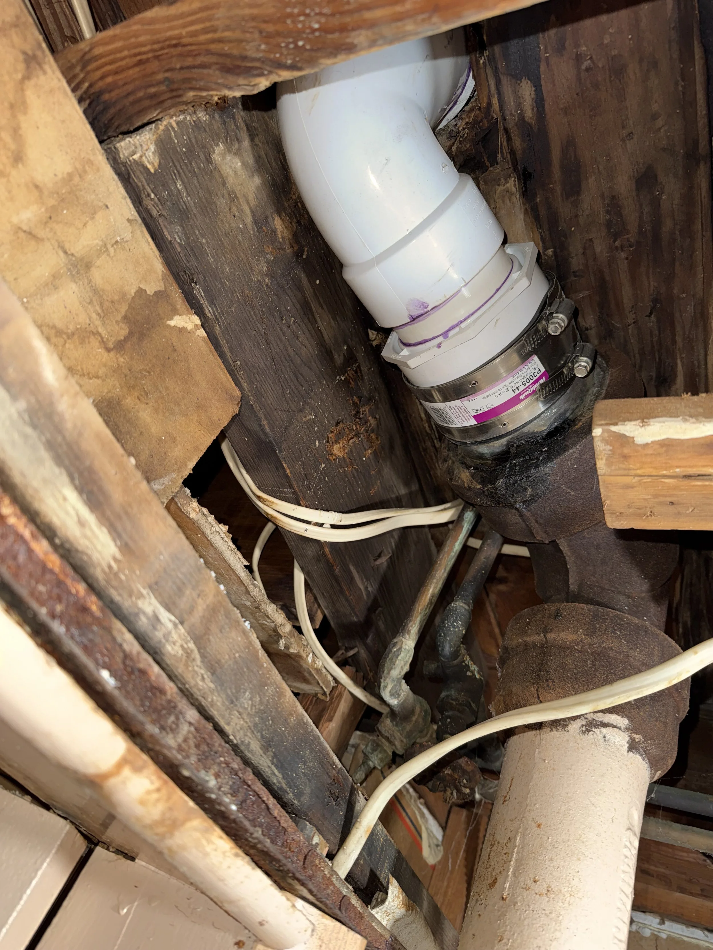 Underneath a kitchen sink showing white PVC pipes, rusted old pipes, and electrical wires attached to a wooden cabinet.