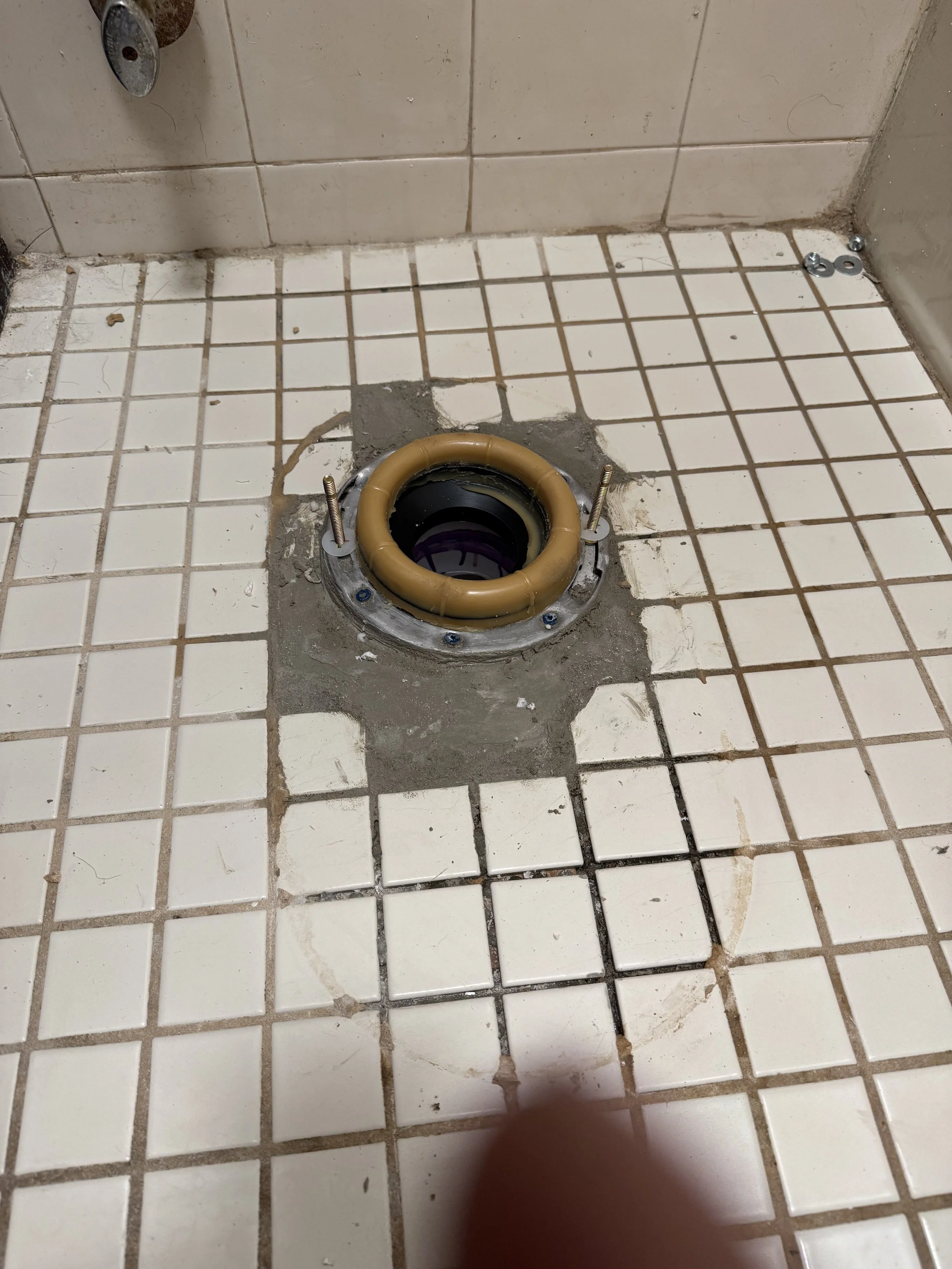 Close-up of a bathroom floor with a toilet flange and a wax ring installed, surrounded by white ceramic tiles, some of which are chipped or dirty.