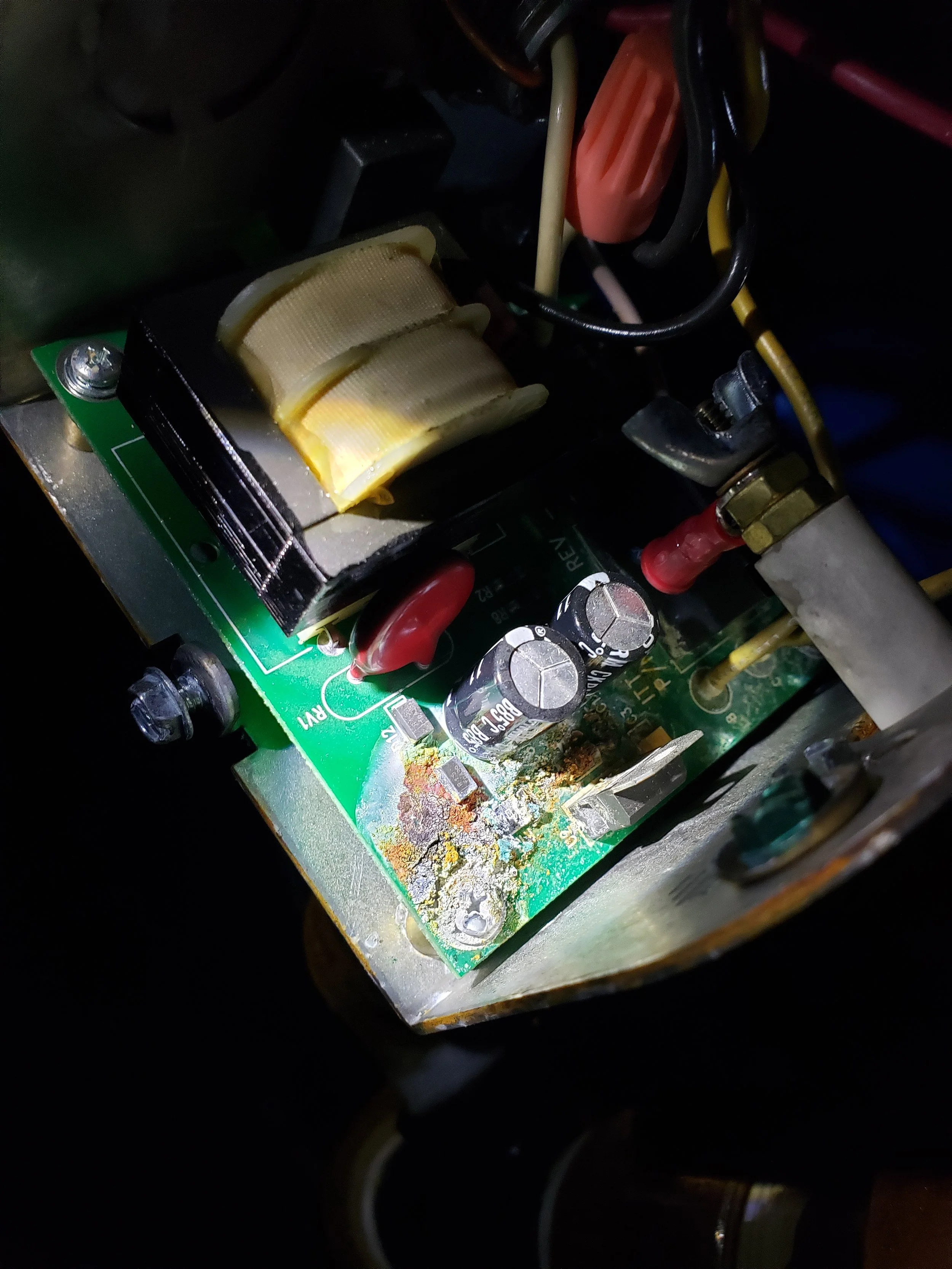 Close-up view of an electronic circuit board with capacitors, wires, and a transformer, showing signs of corrosion and residue.