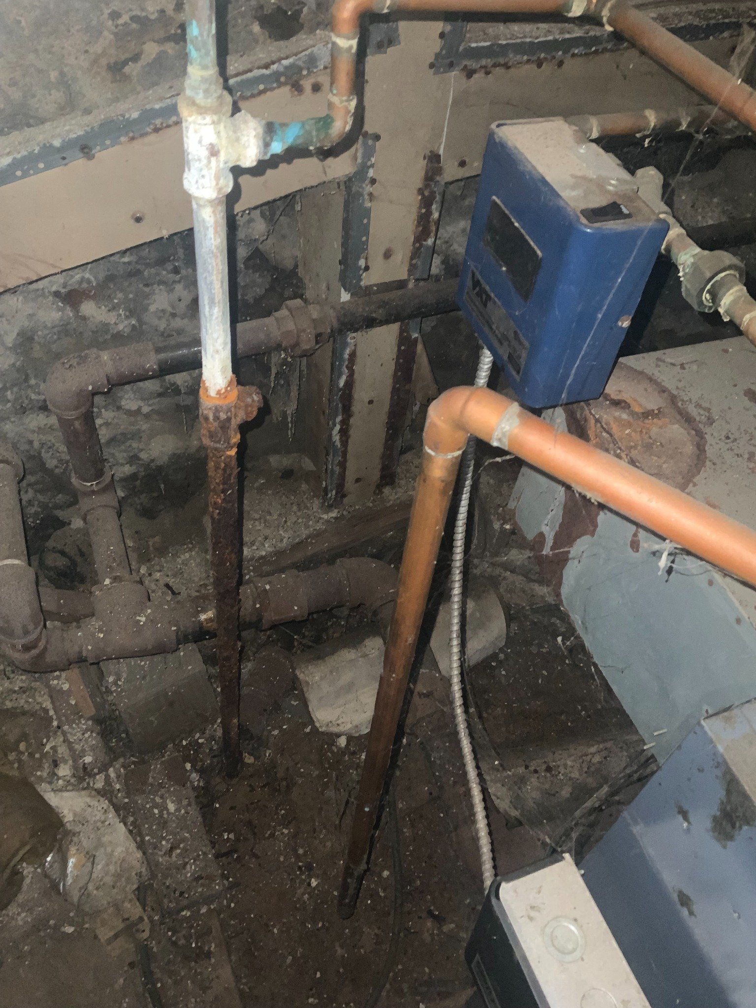 Close-up of an industrial plumbing system with rusted pipes, a blue digital meter, and electrical wiring in a construction or maintenance setting.