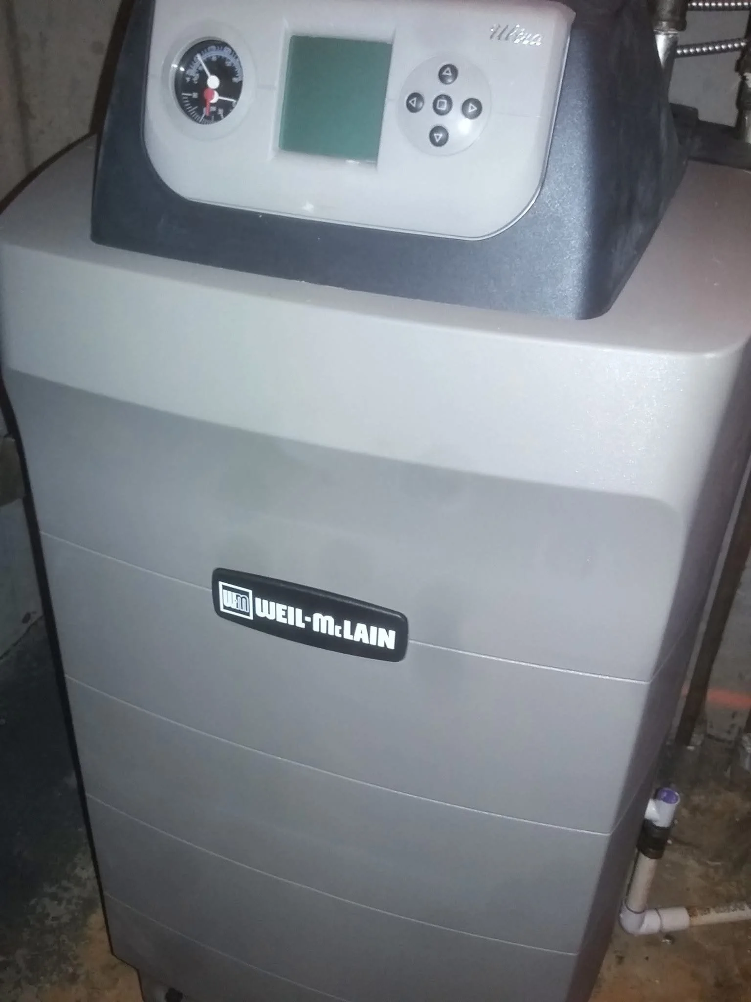 A large, gray Weil-McLain boiler with a black control panel on top, placed on a concrete floor near a wall and some pipes.