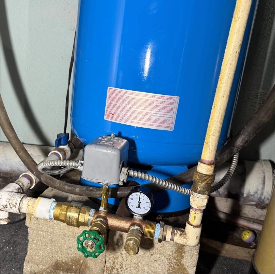 Plumbing system with a blue tank, pressure gauge, brass, copper, and PVC pipes, and a green valve handle.